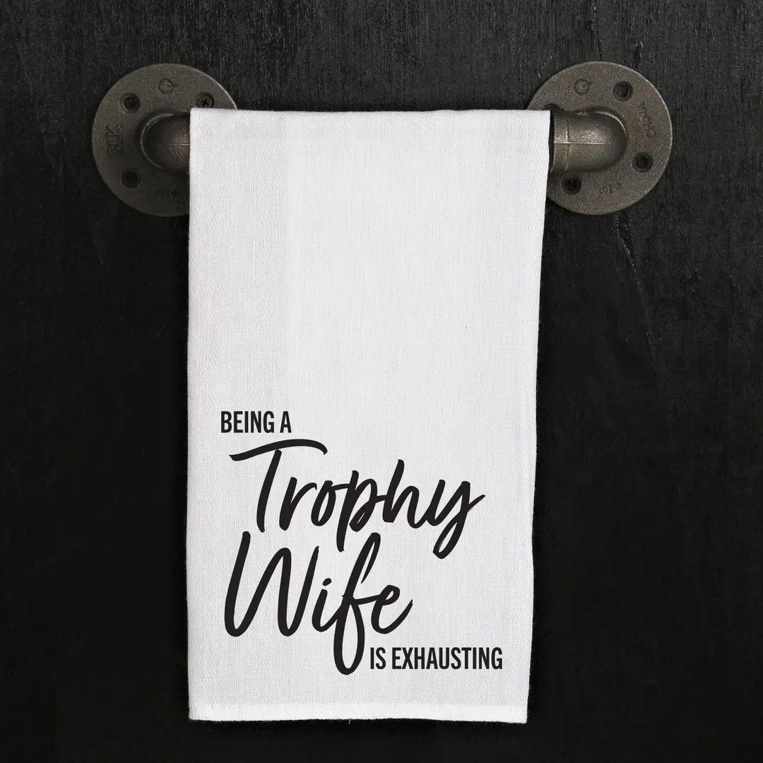 White towel on a dark rack reading “Being a Trophy Wife is Exhausting.”