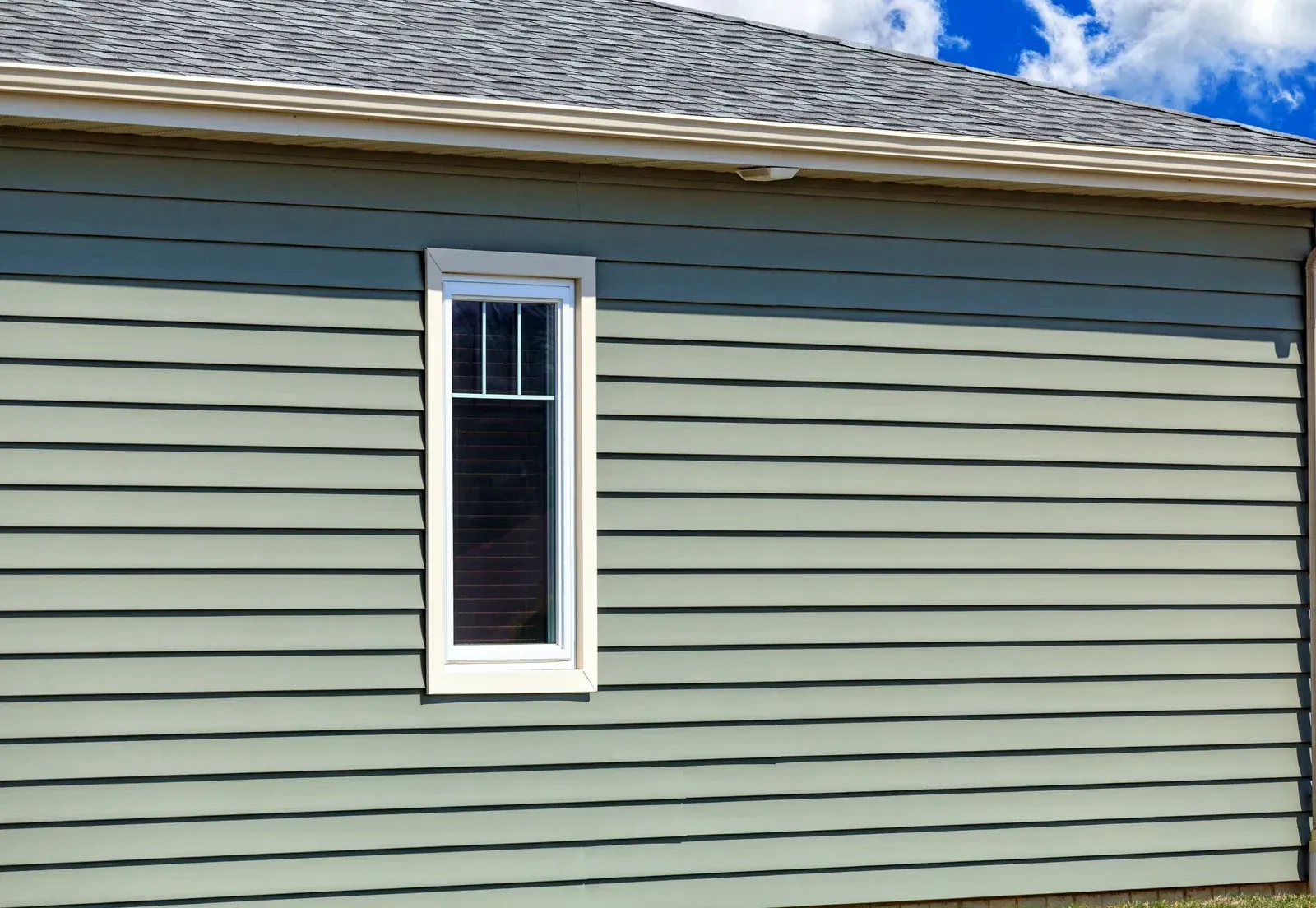 Side of a house with light green siding, a tall narrow window, and gray roof.
