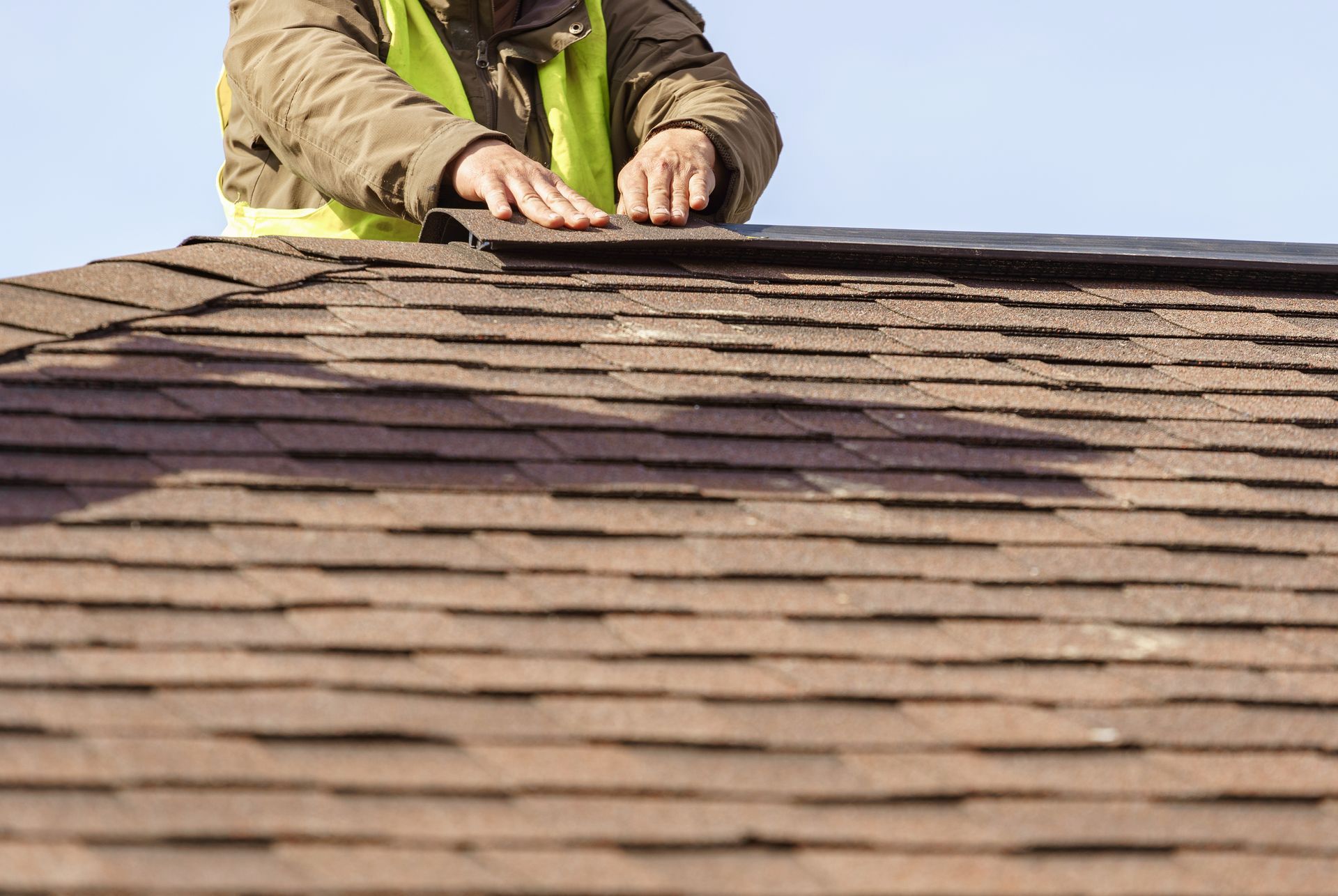 Worker inspecting a shingled roof from above, hands near the ridge line