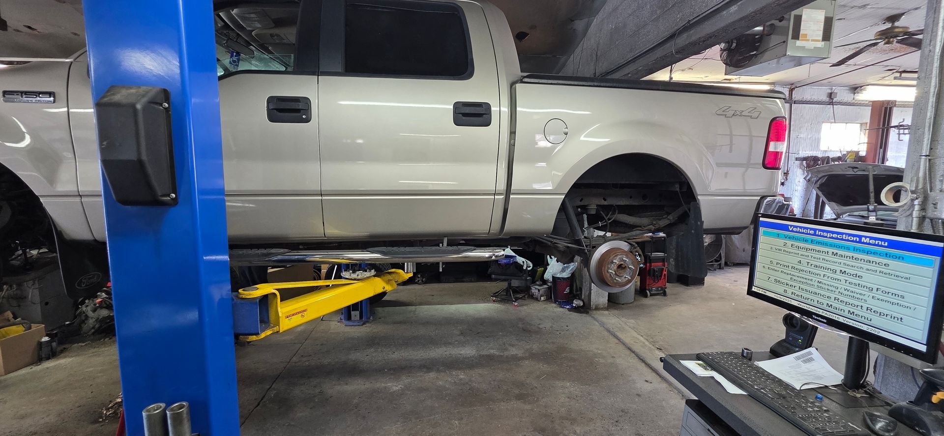 A light-colored pickup truck is raised on a blue hydraulic lift inside a garage, with a computer screen visible nearby.