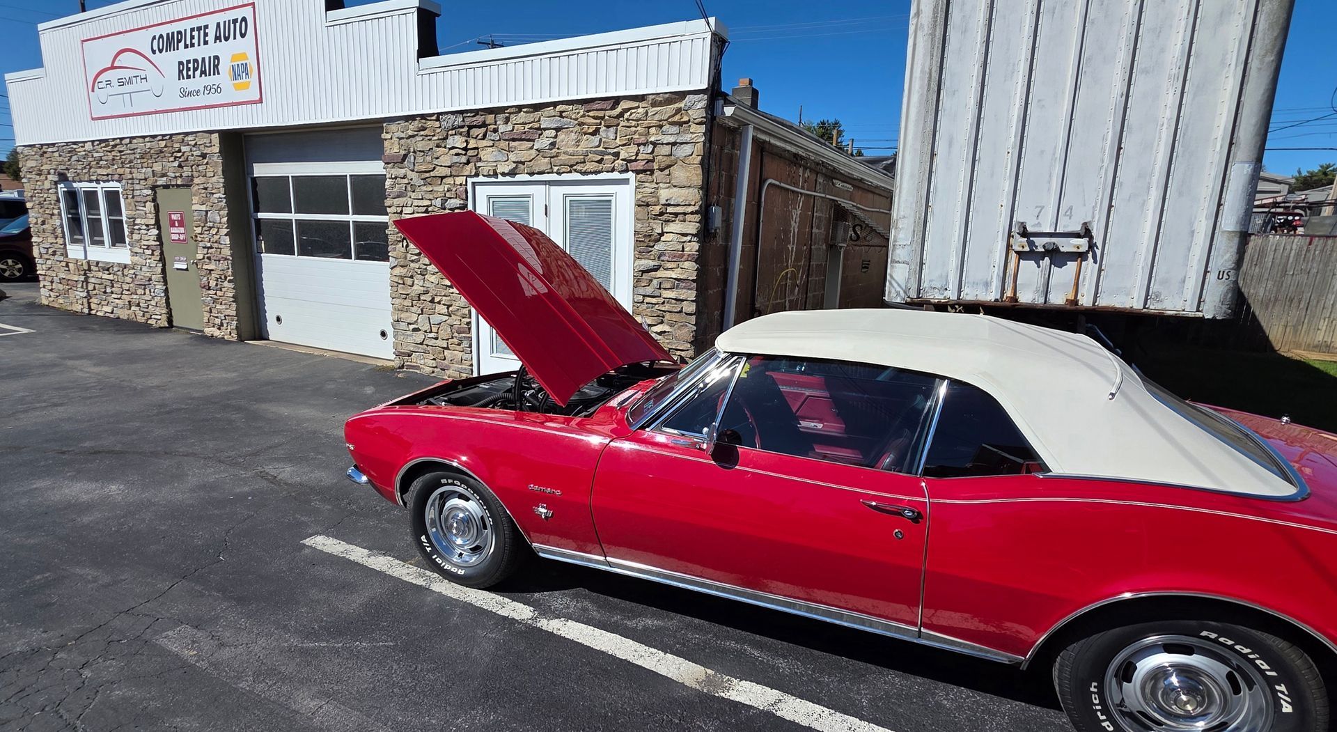 A classic red convertible with its hood up parked in front of a stone-faced automotive shop on a sunny day.