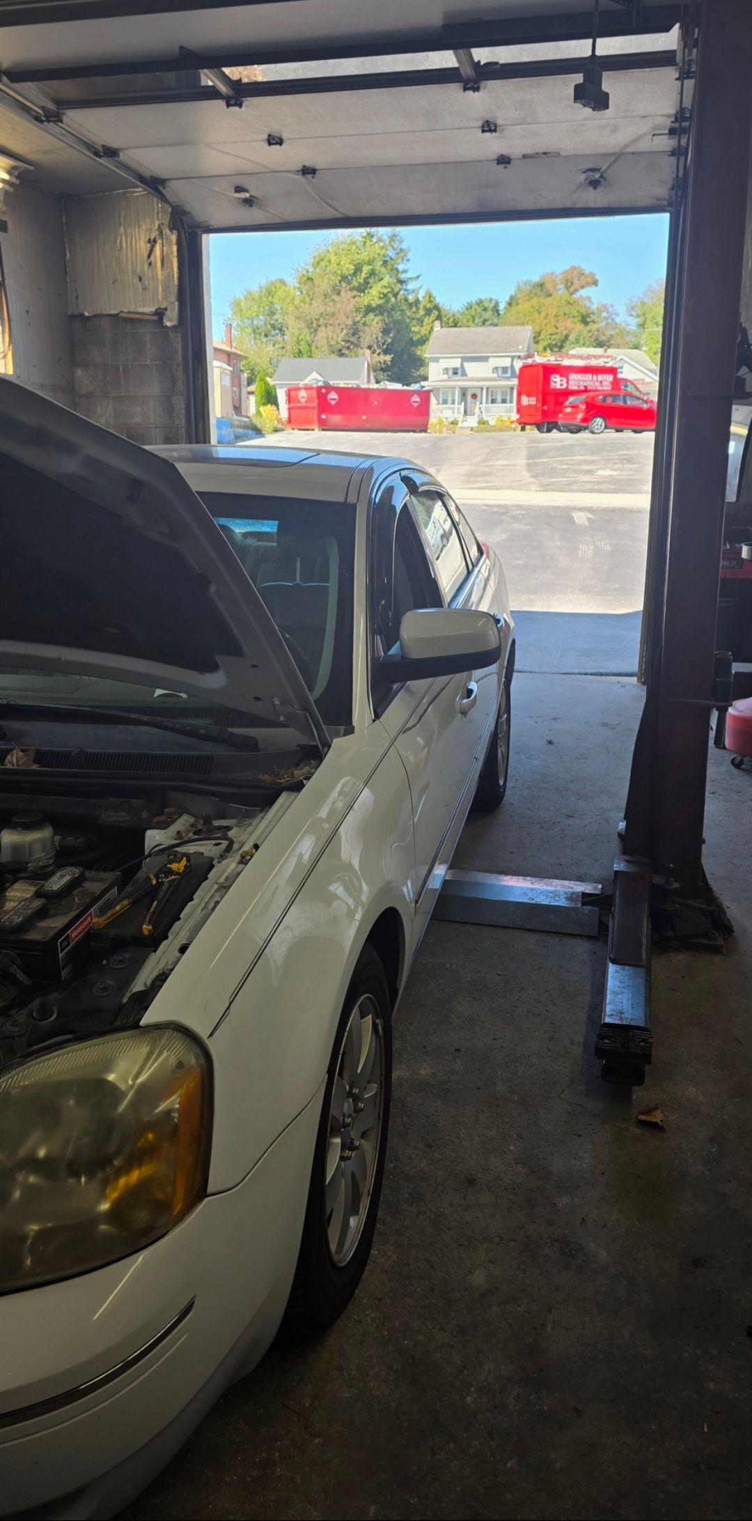 A white car with an open hood is parked inside a garage bay, viewed from the side looking out toward a sunny street.