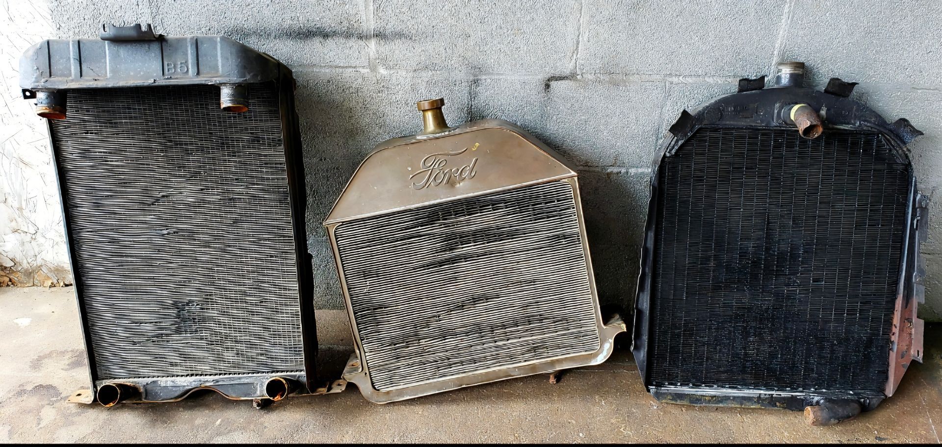 Three vintage vehicle radiators stand side-by-side against a concrete wall.