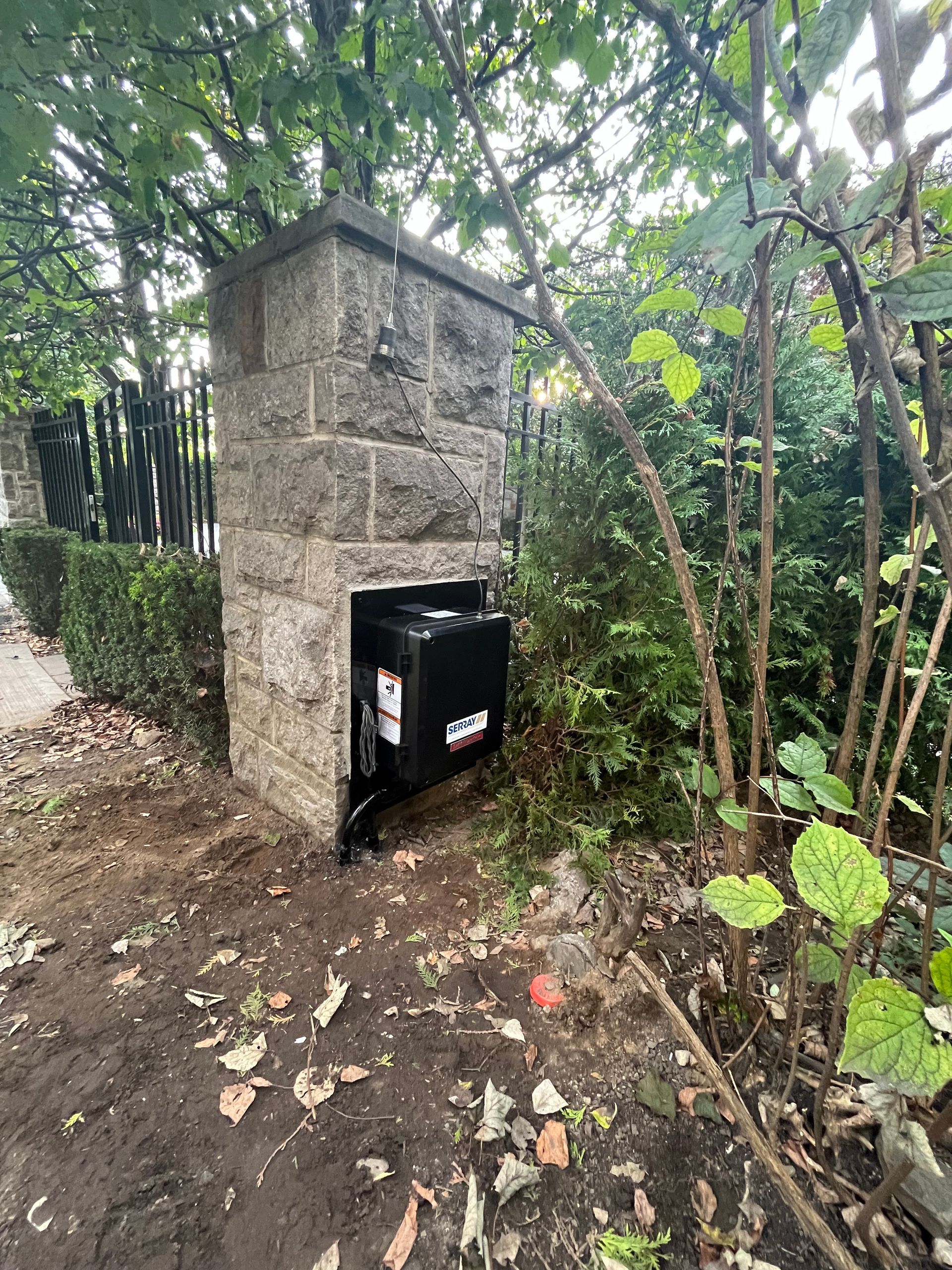 Un pilier en briques avec une boîte noire à l'intérieur est entouré d'arbres et de buissons.