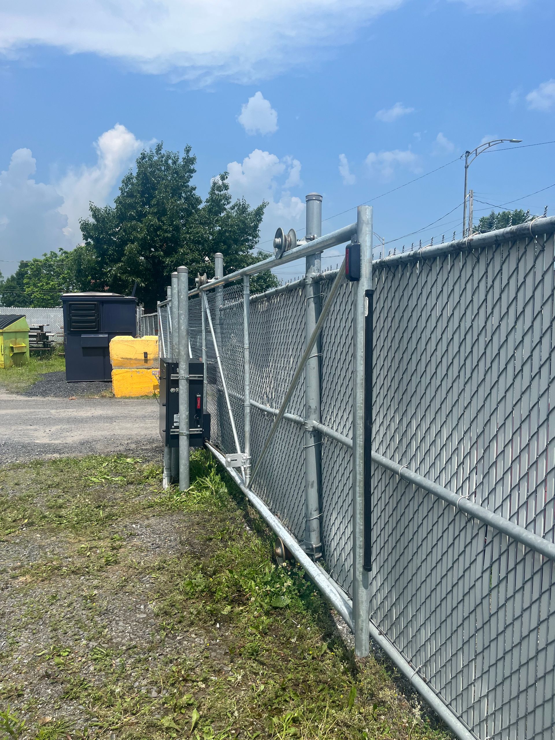 Une clôture à mailles losangées avec un portail coulissant et une benne à ordures jaune en arrière-plan.