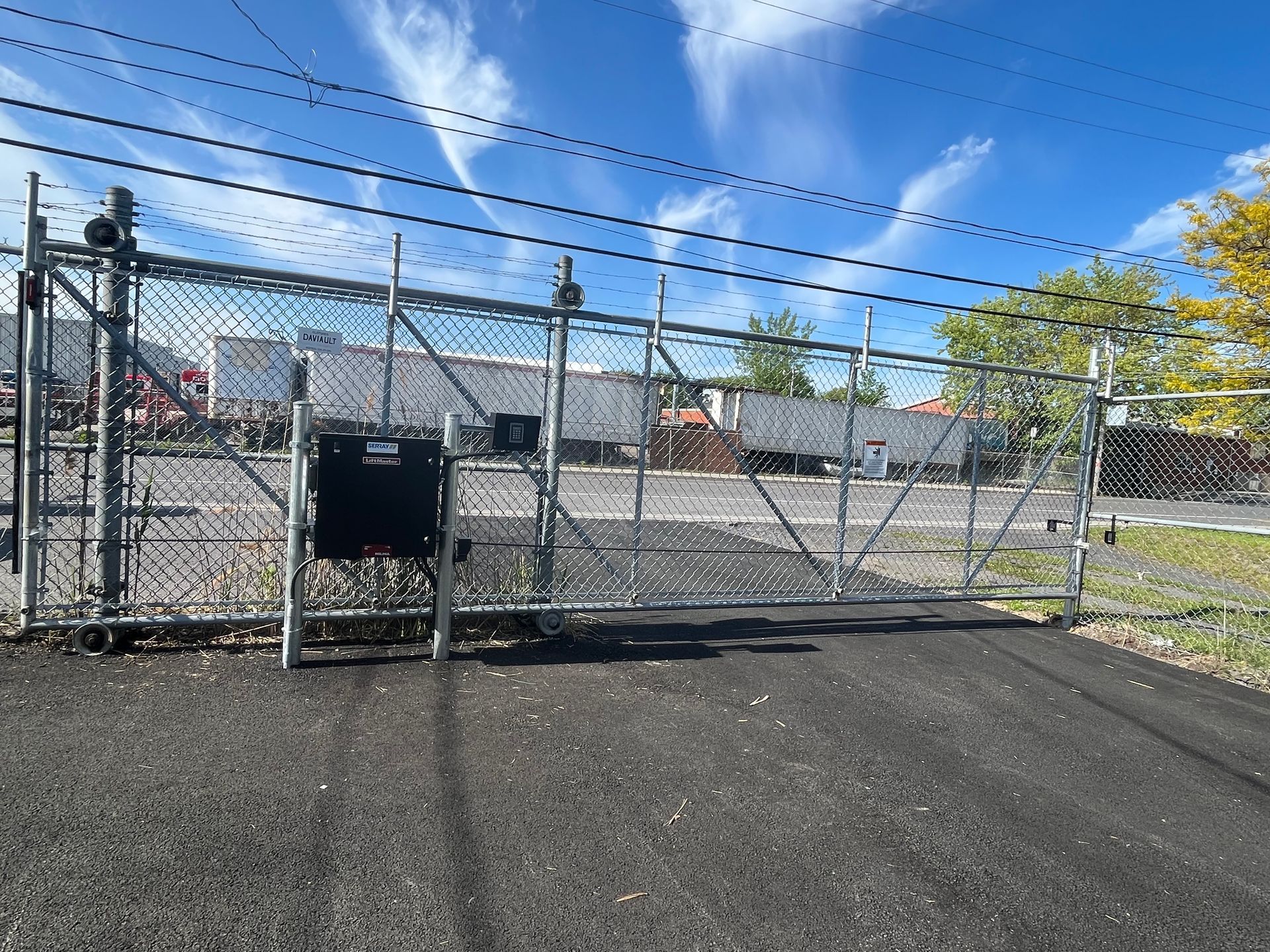 Une clôture à mailles losangées avec un portail coulissant devant un parking.