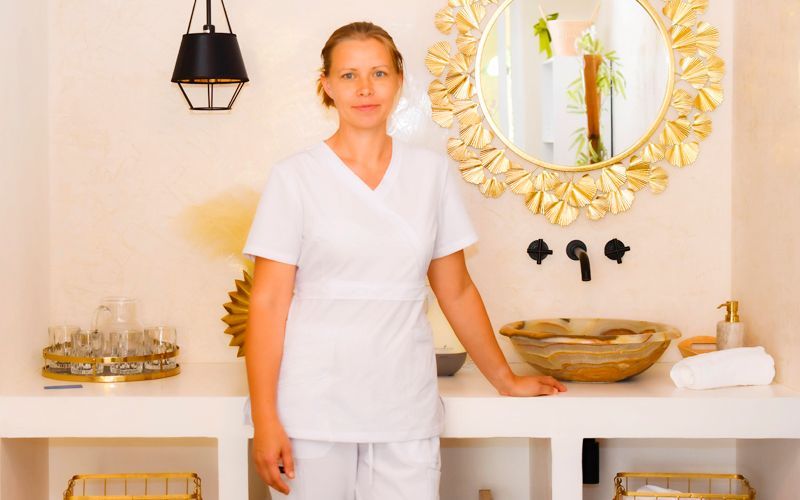 A woman in a white scrub top is standing next to a sink in a bathroom.