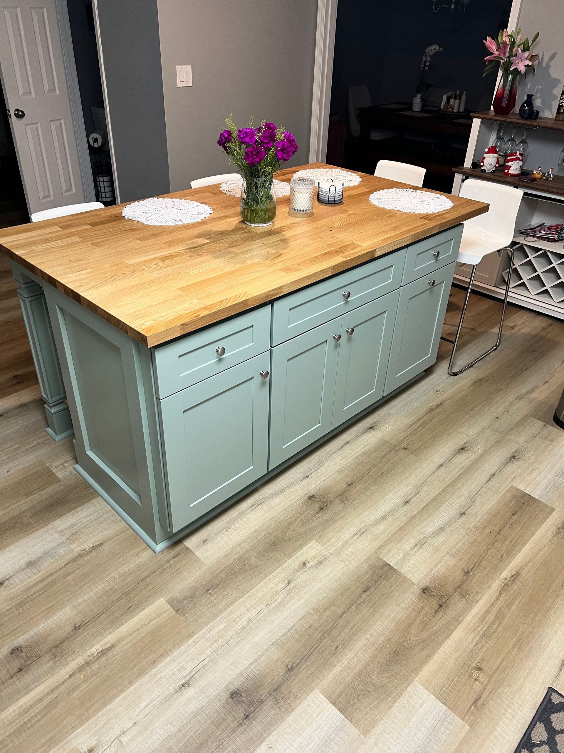 Kitchen island with a light wood top, teal cabinets, flowers, and white chairs.