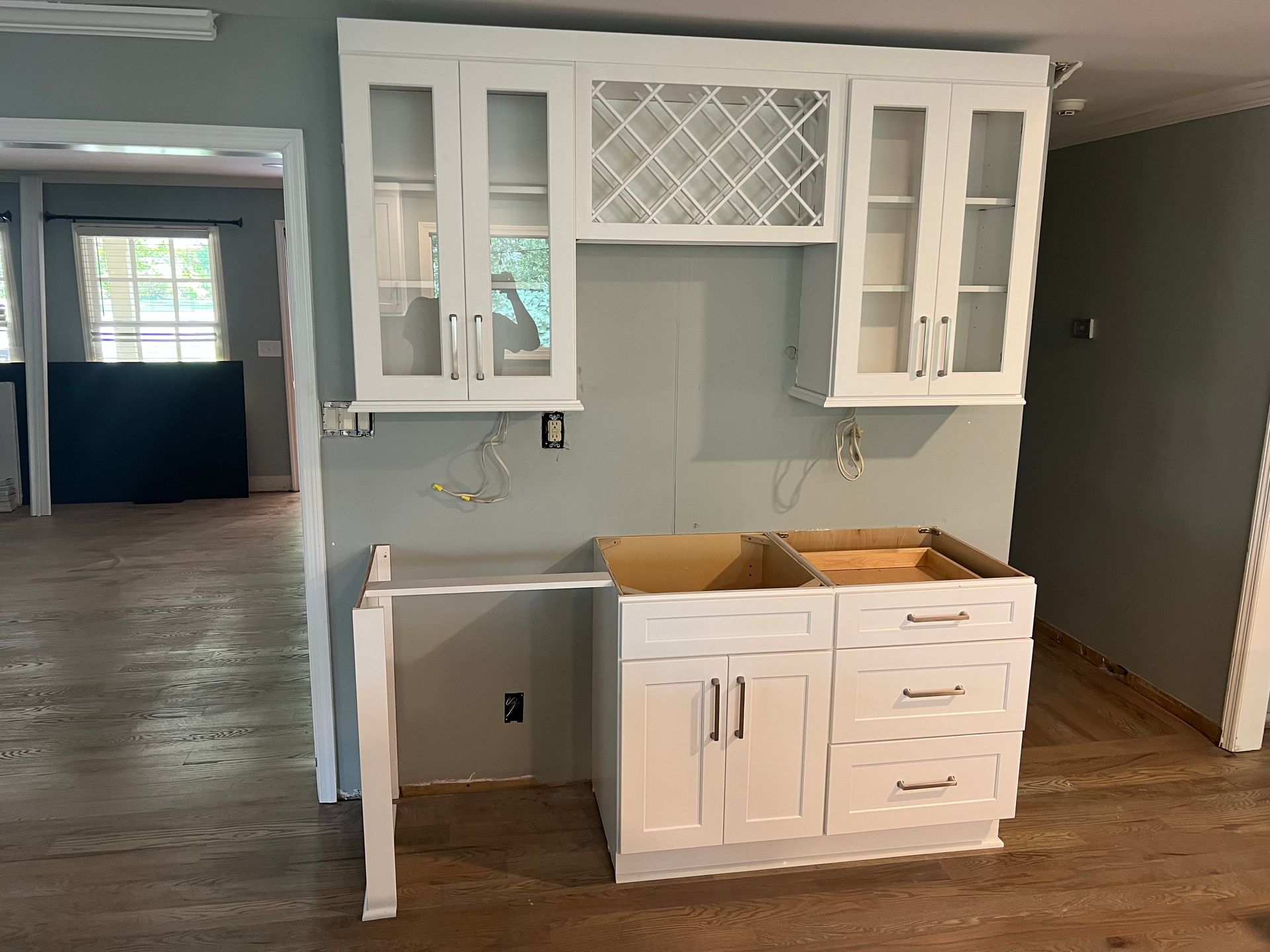 White kitchen cabinets installed against a gray wall; incomplete; wood floor.