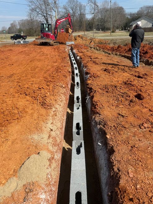 Utility trench with a black conduit being laid beside an orange dirt road, with a backhoe and worker nearby