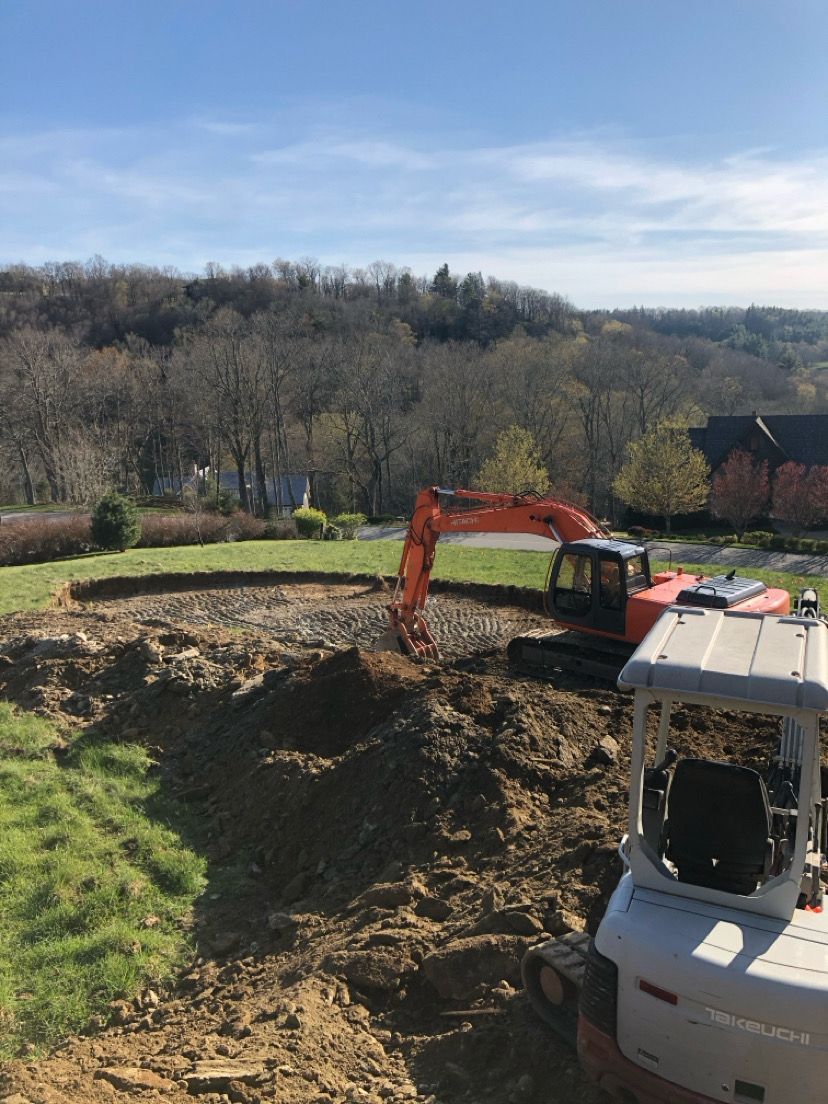 Excavator on a dirt excavation site beside a white truck, with grassy hills and leafless trees in the background
