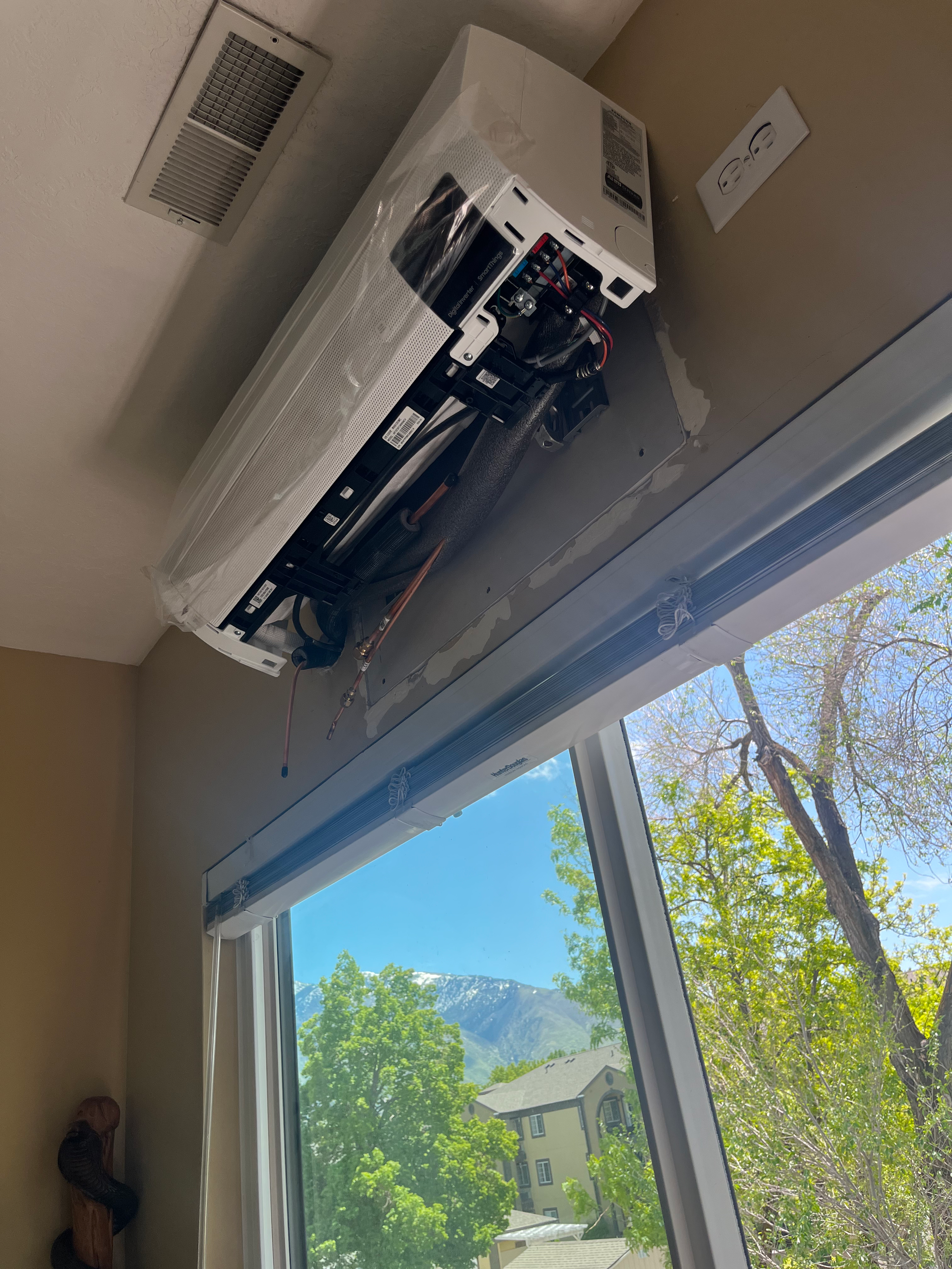 An air conditioning unit is detached from the wall, above a window with a mountain view.