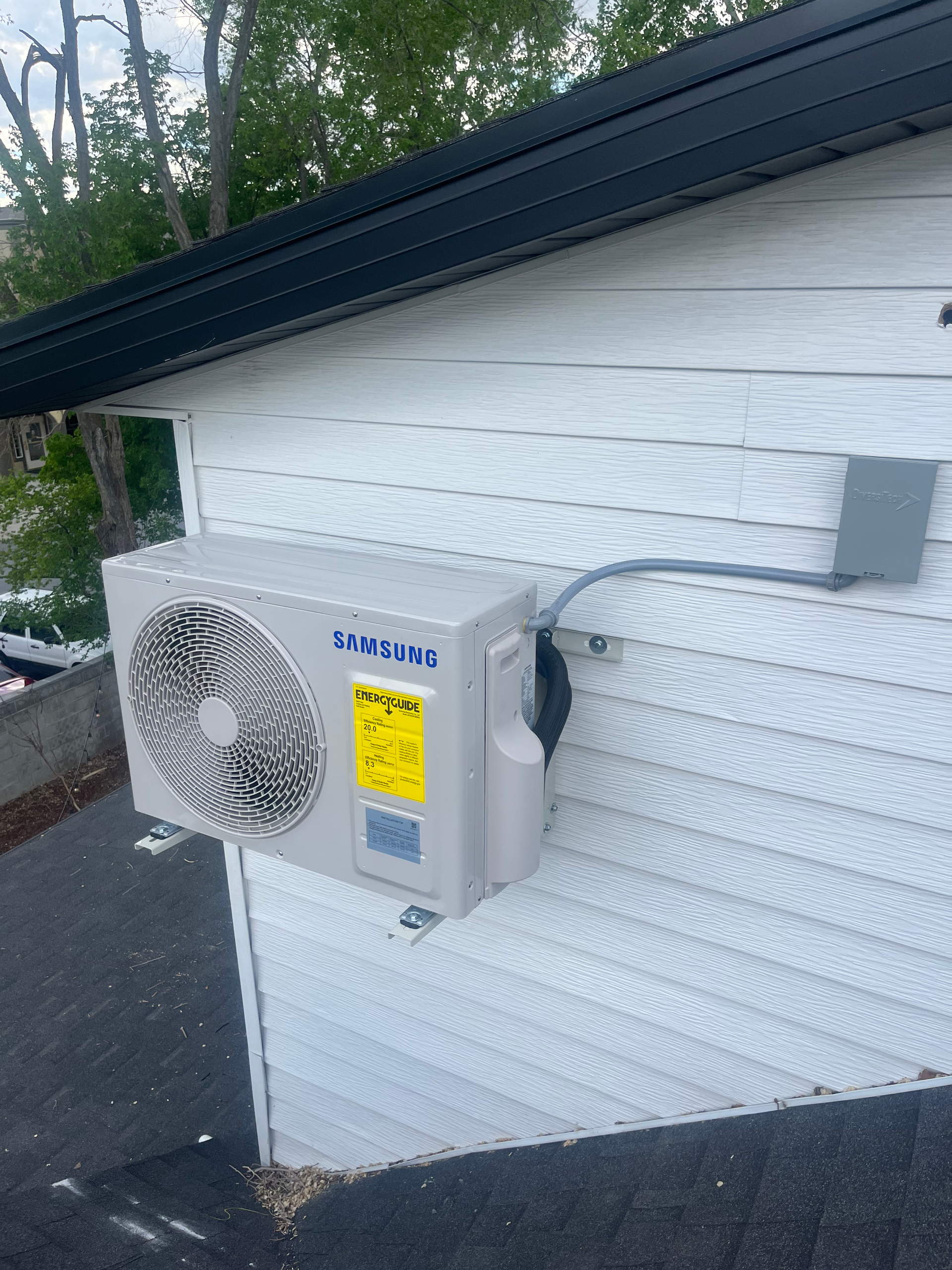 A Samsung air conditioning unit mounted on a white house exterior near a black roof.