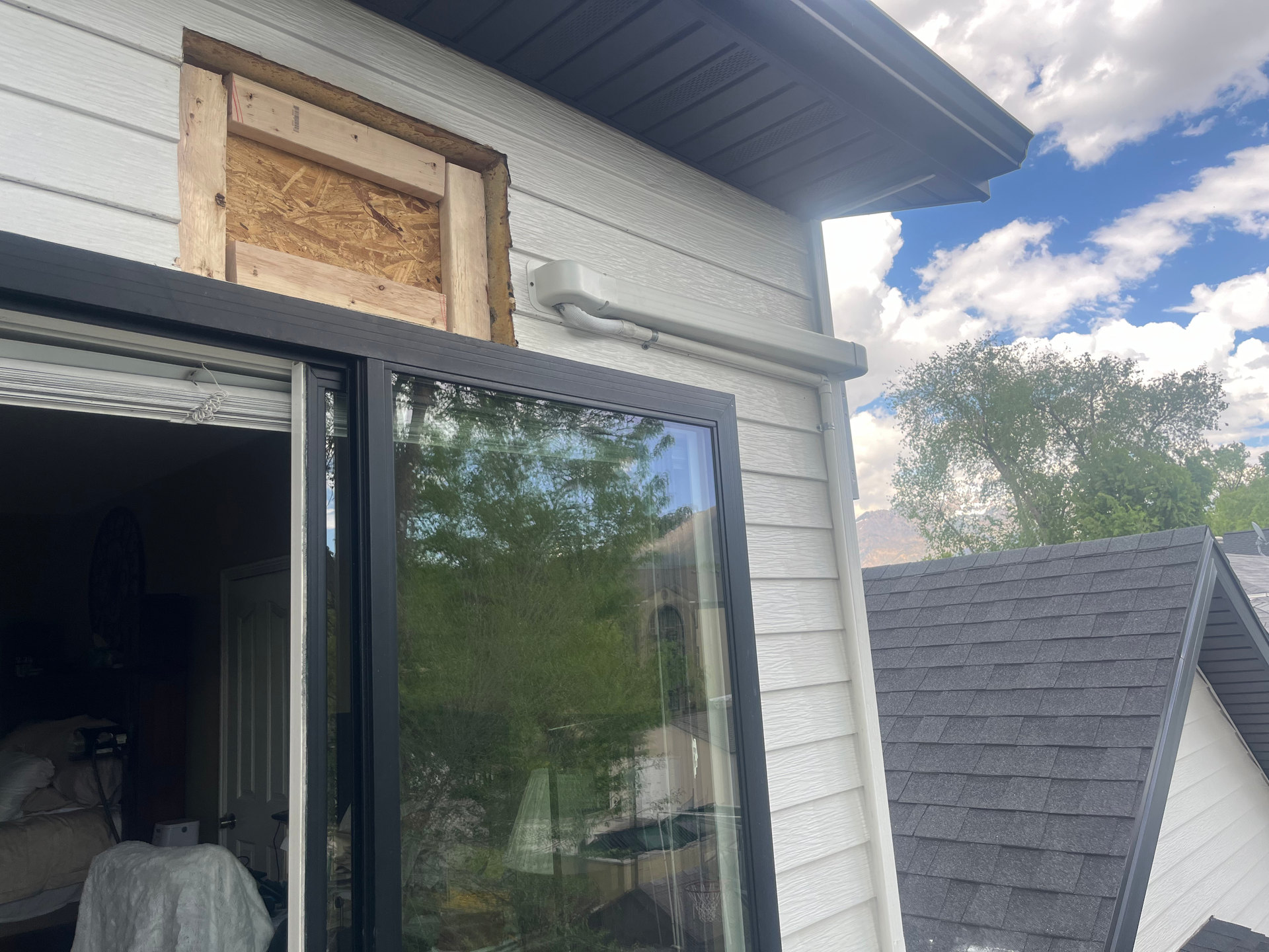 House exterior with sliding glass door and boarded-up window. White siding, black trim, blue sky.