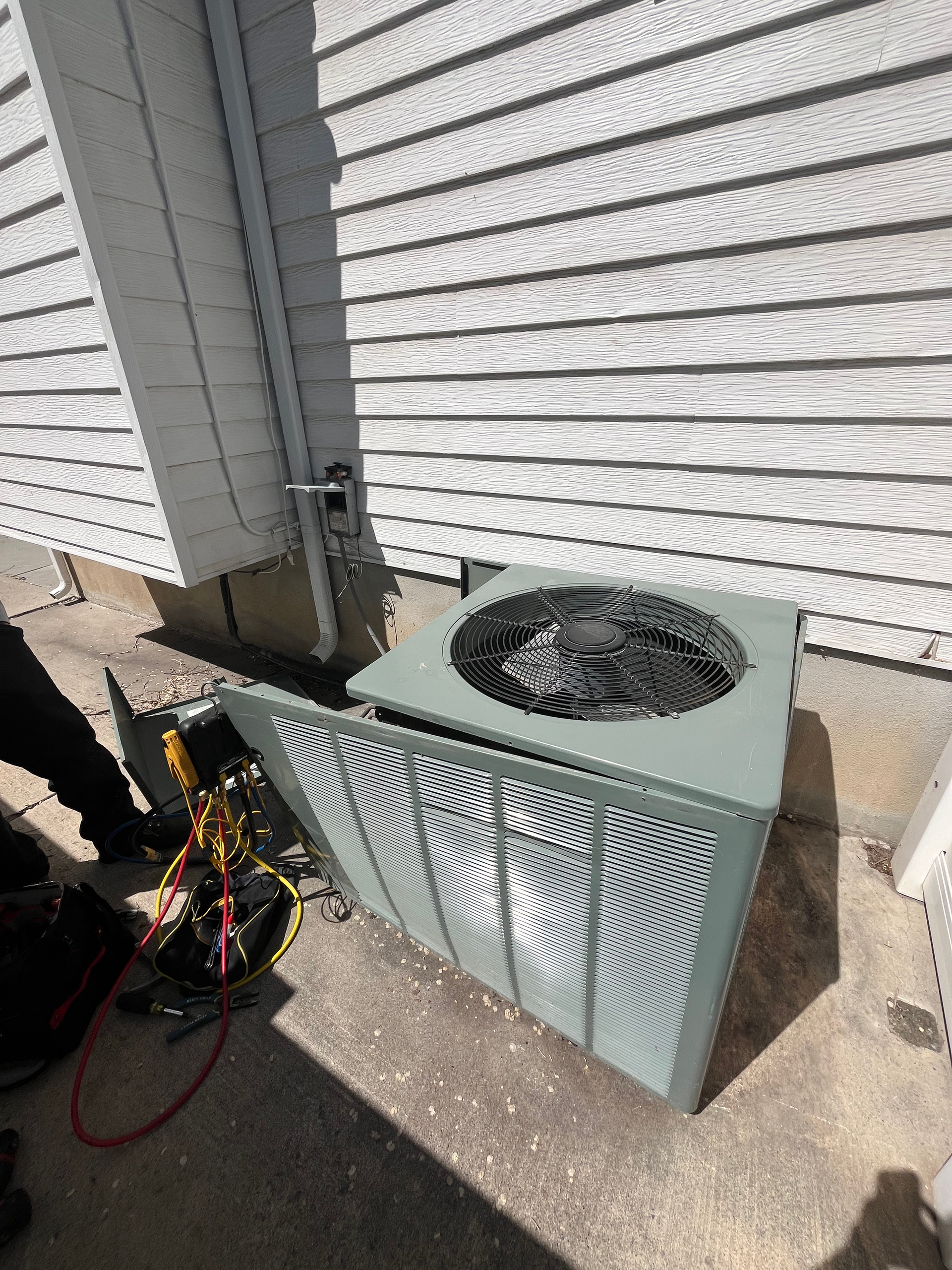 HVAC unit with side panel open; tools visible on ground, against a white siding wall.