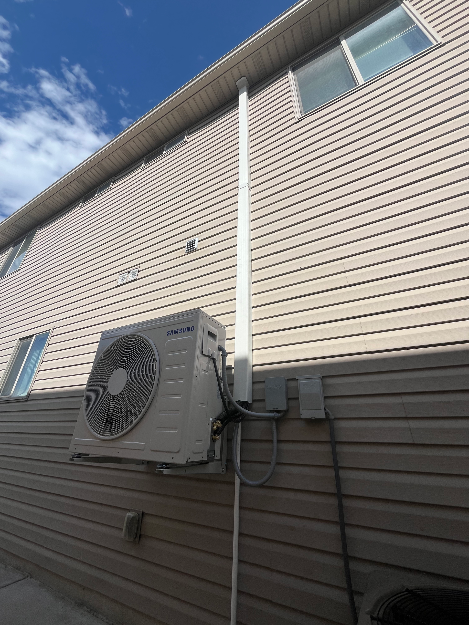 Air conditioning unit mounted on a light brown siding wall with a white pipe running up, blue sky in background.