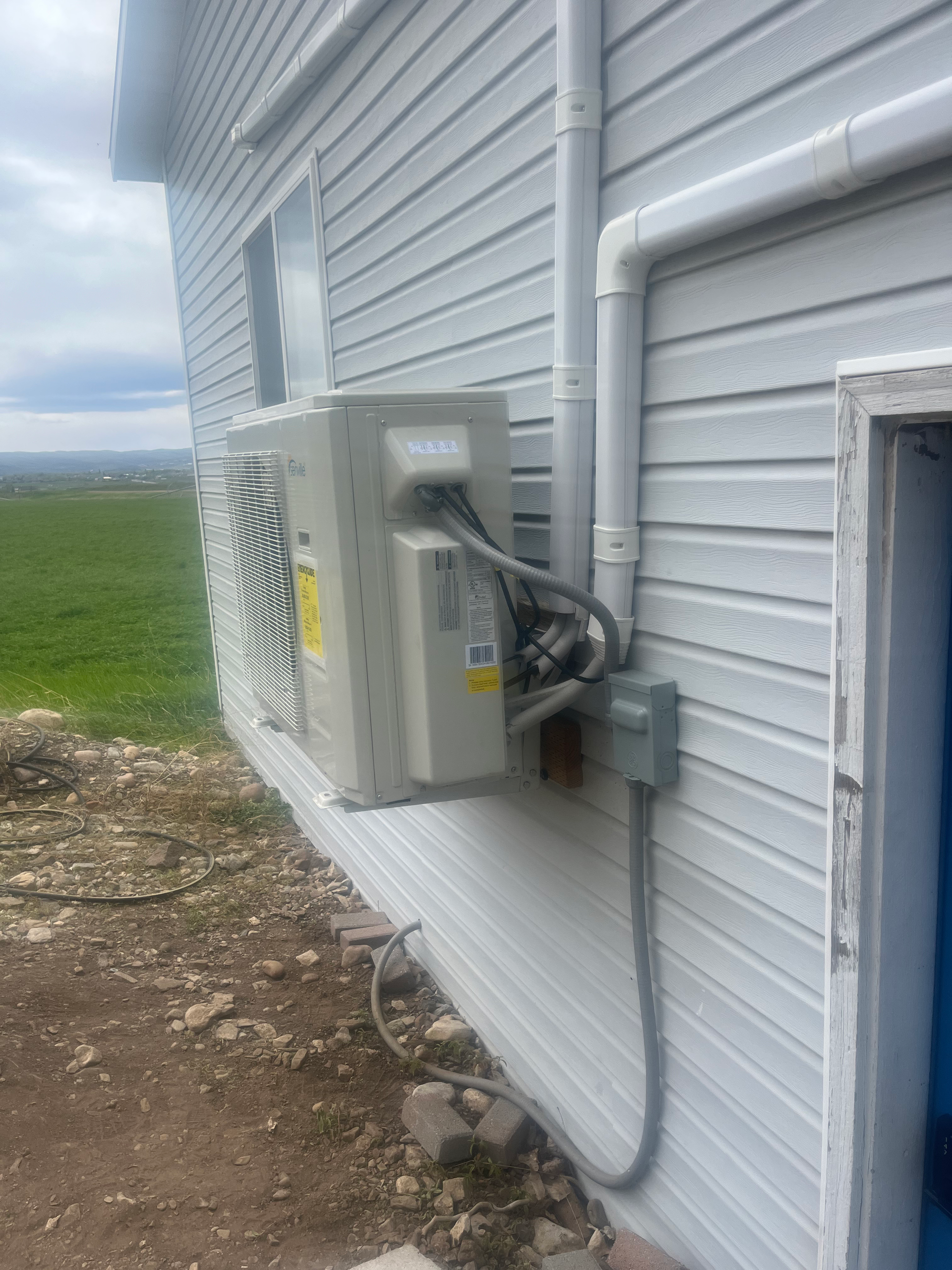 Outdoor air conditioning unit mounted on a light gray house wall with gray electrical conduit.