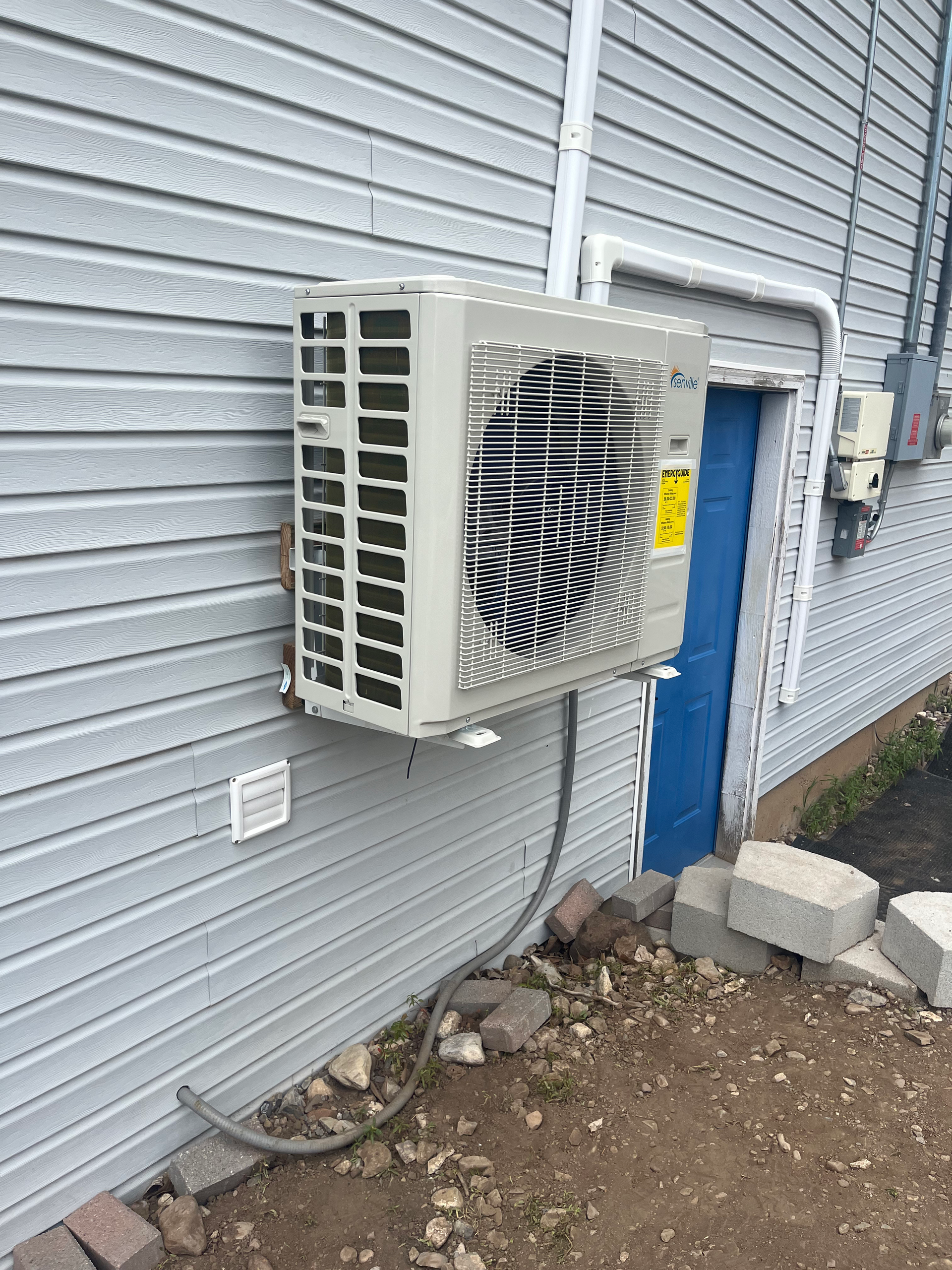 An air conditioning unit mounted on a gray, clapboard house wall next to a blue door.