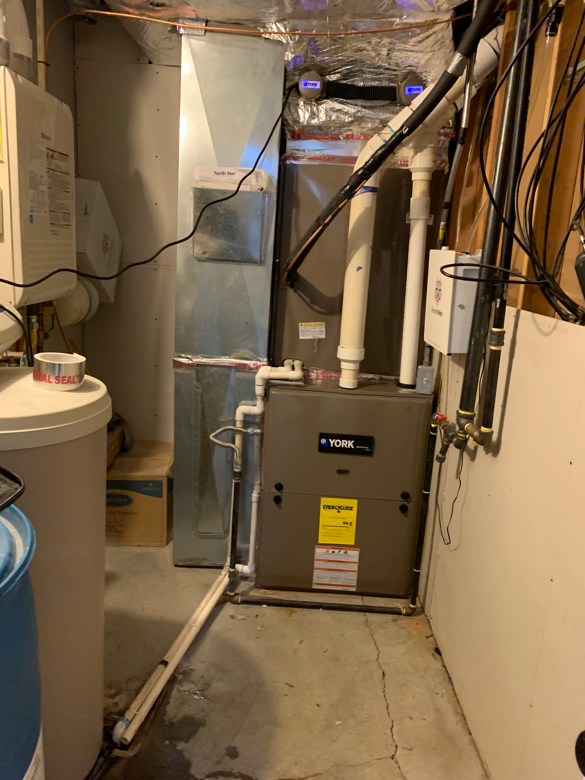 Furnace and ductwork in a basement setting. Gray and silver components, white pipes, water tank and electrical panel.