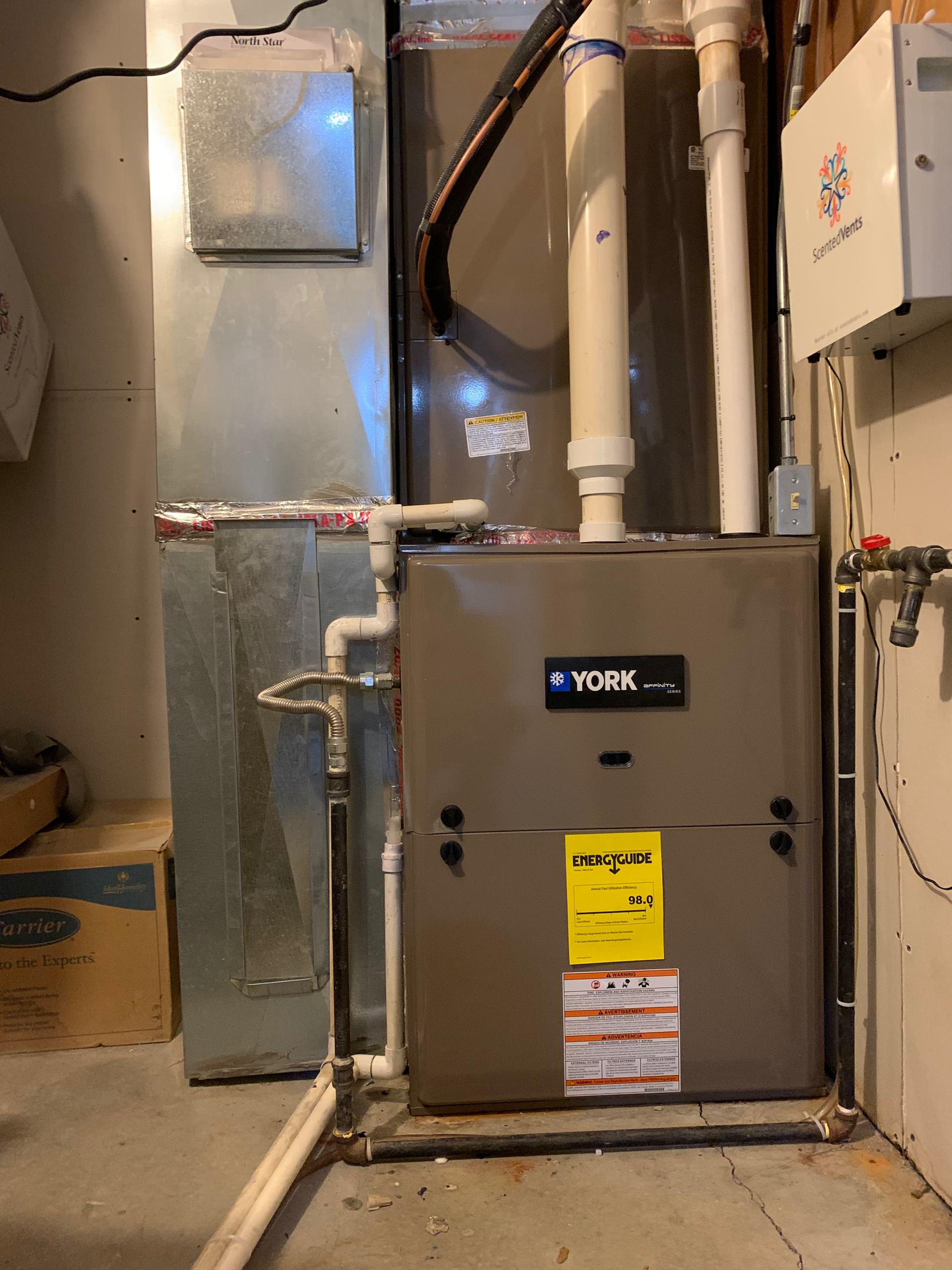 A furnace with ductwork in a basement setting. The unit is beige, with a yellow energy guide label.