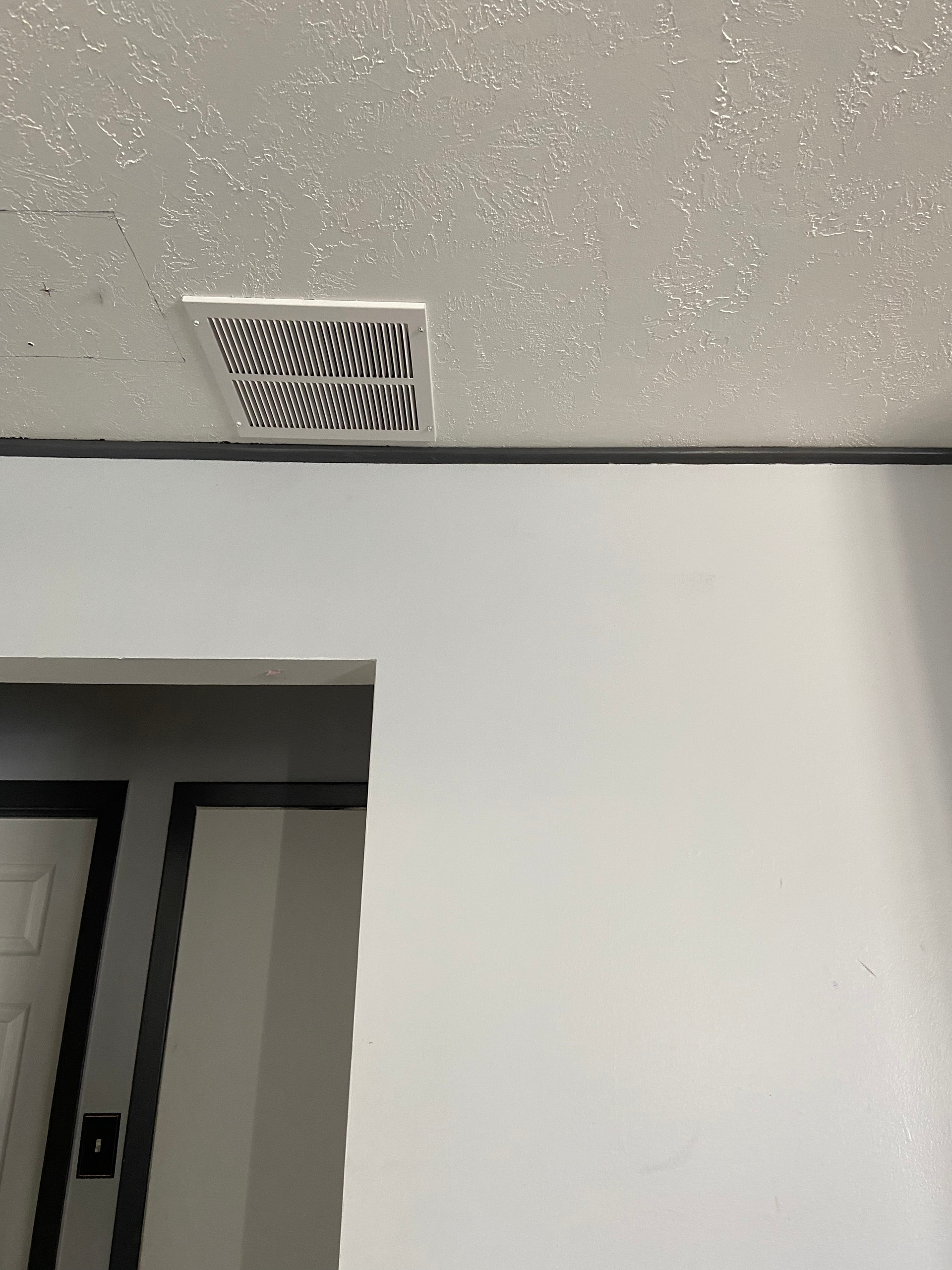 Ventilation grate in a textured ceiling above a doorway. White wall and trim, black outline on the door.