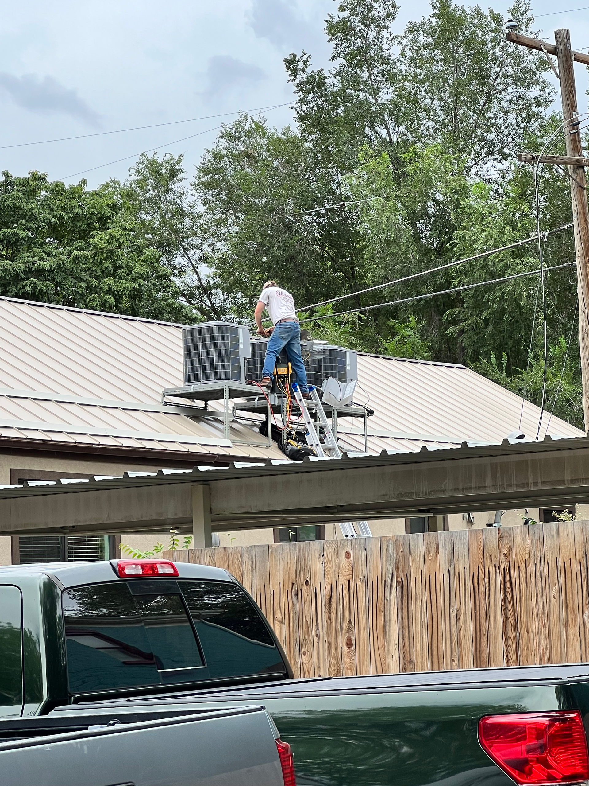 Person on ladder near AC units on a roof, working. Green trees, power line, and parked truck in the background.