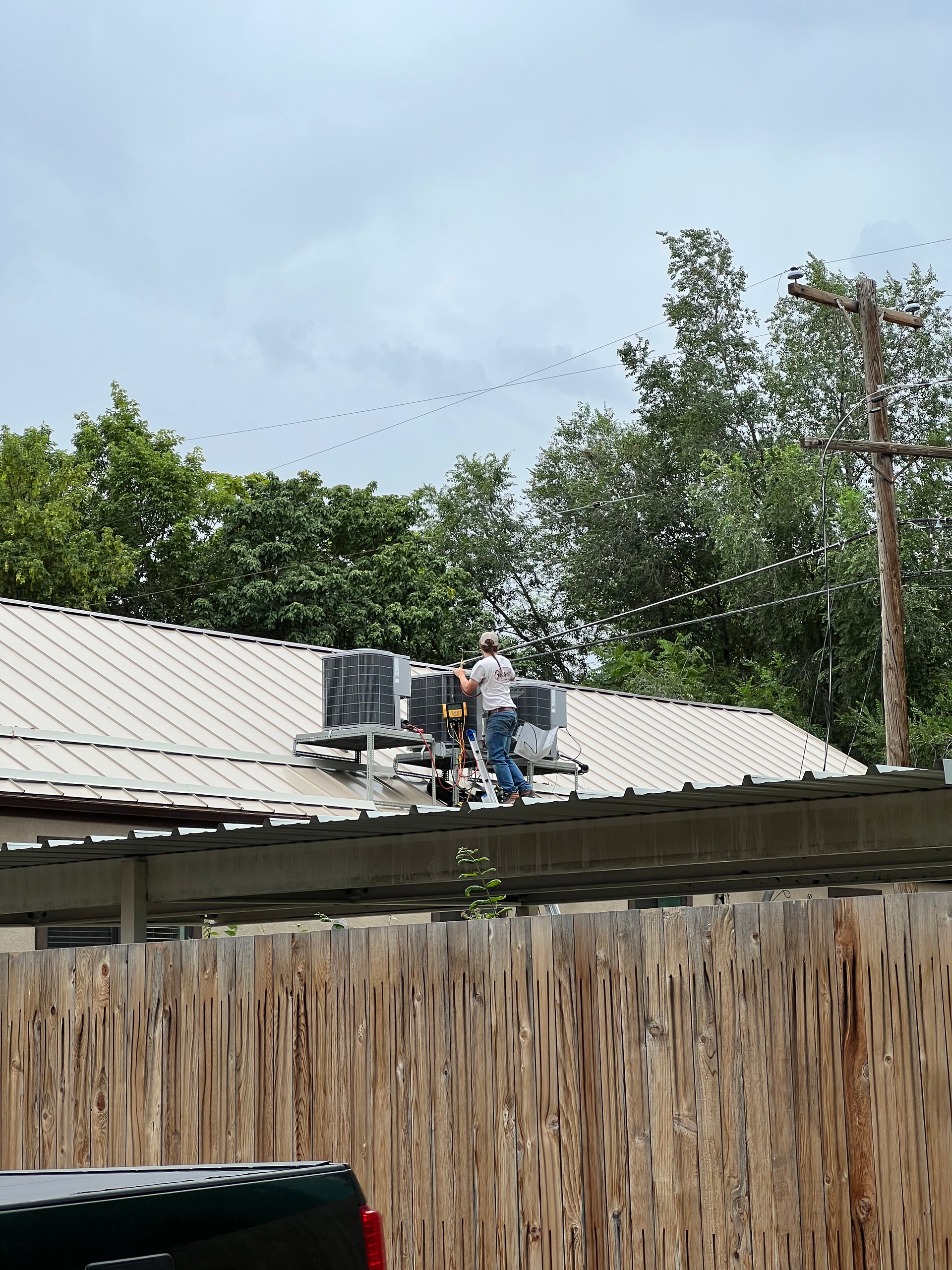 Man working on rooftop AC units, surrounded by trees and flock of birds. Wooden fence in foreground.