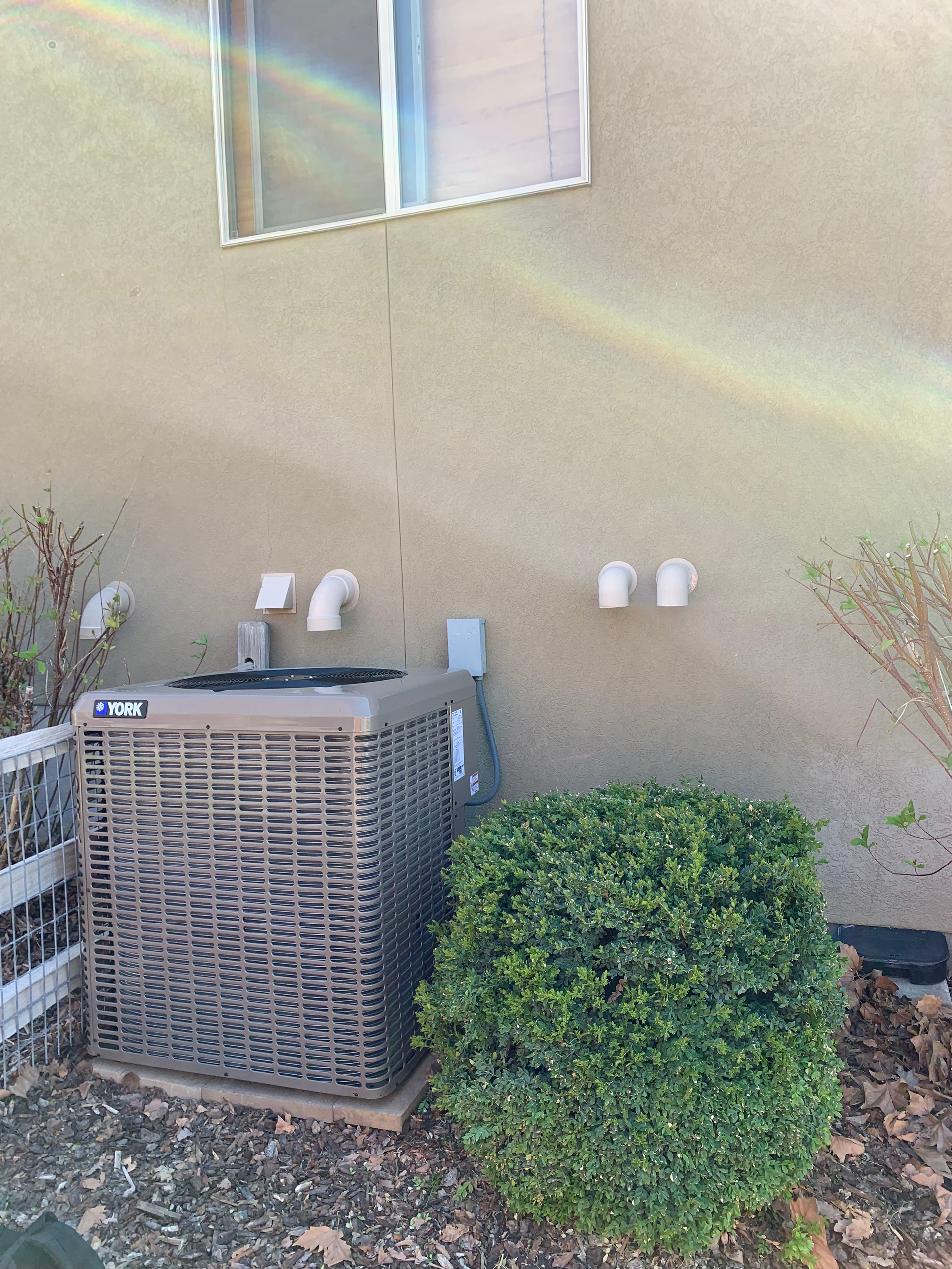 Air conditioner unit outside a tan stucco building with a green bush.