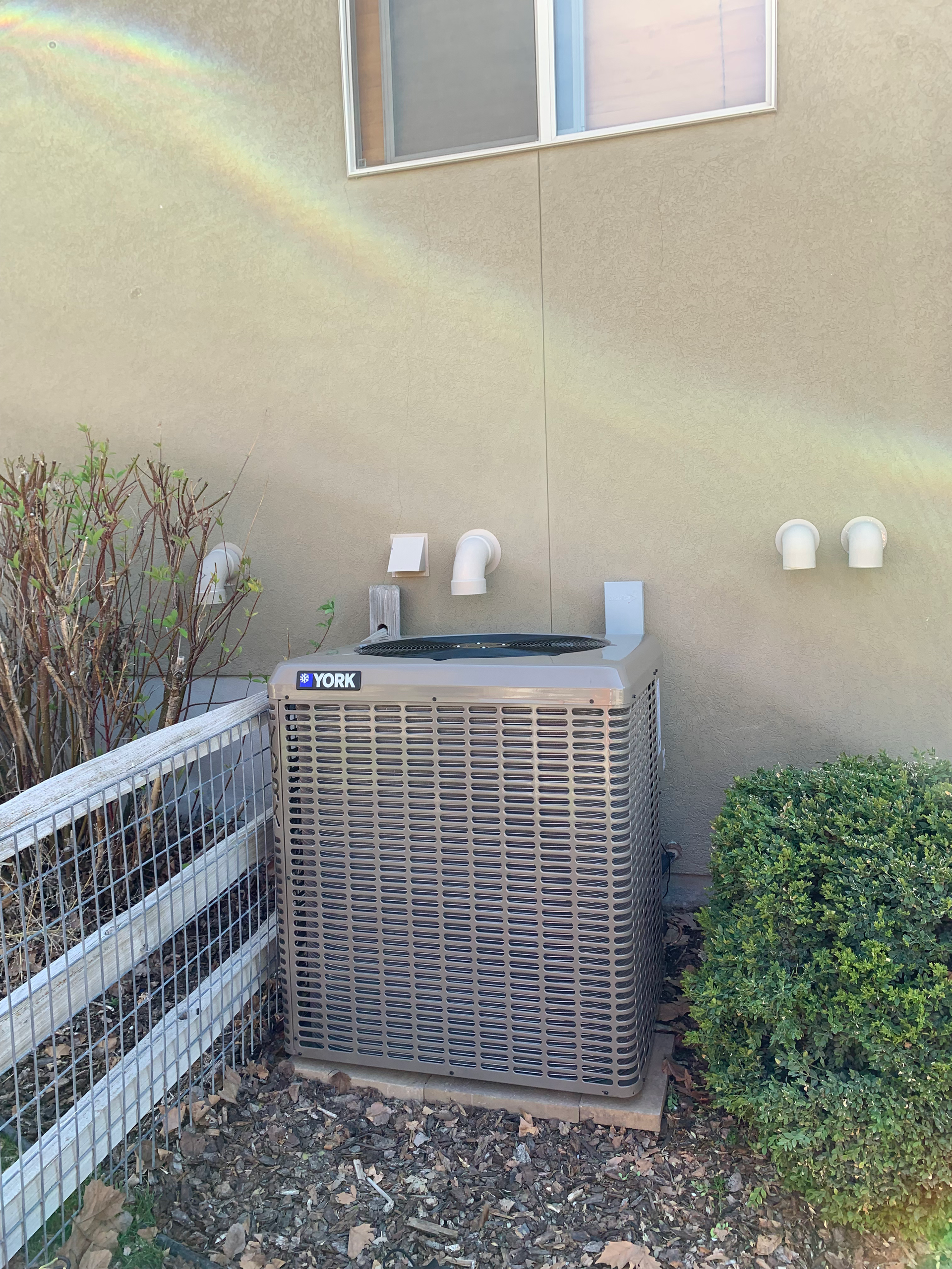 An outdoor air conditioning unit next to a building with a fence and a window.