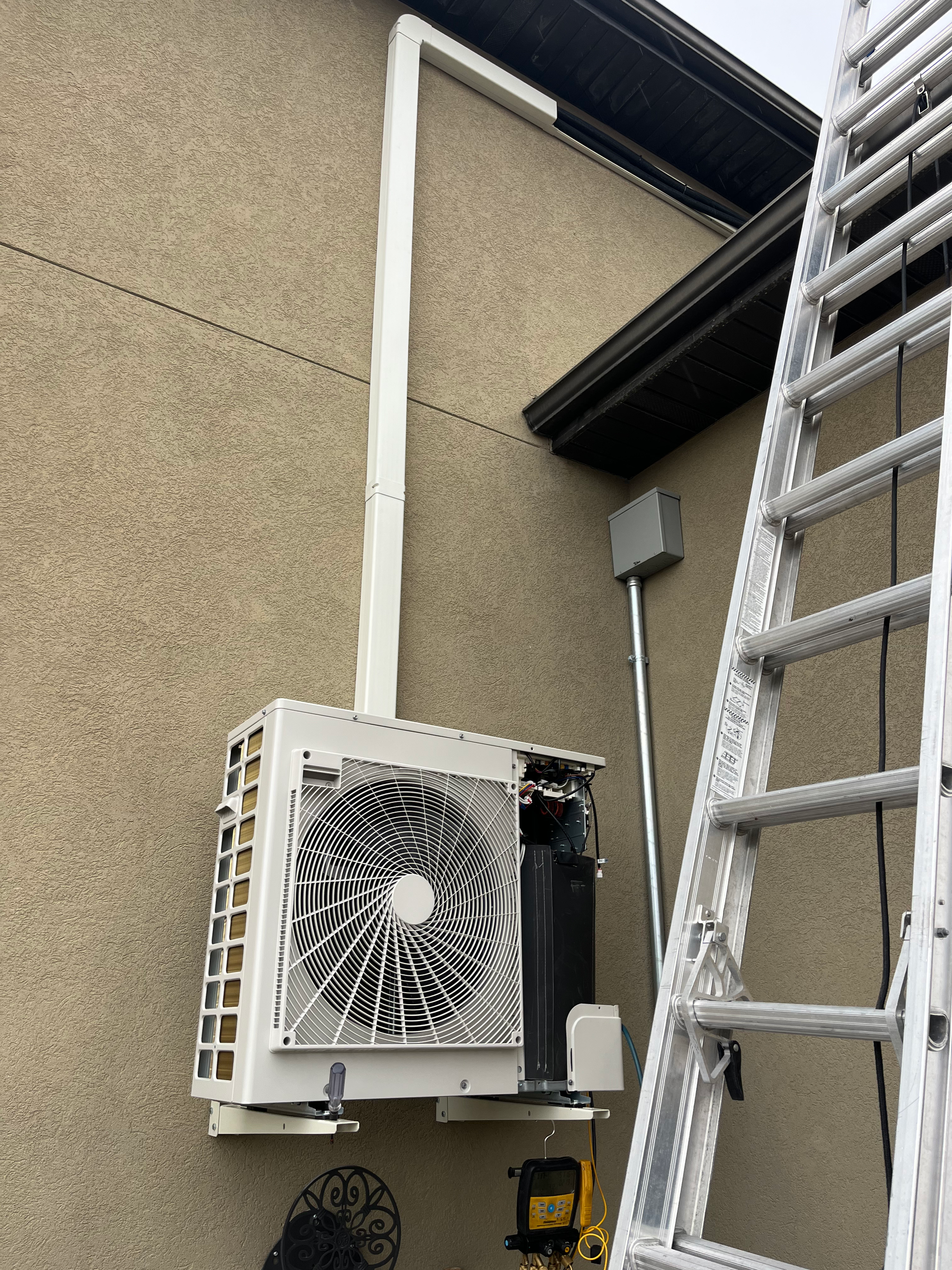 White heat pump mounted on tan stucco wall, with conduit and ladder.