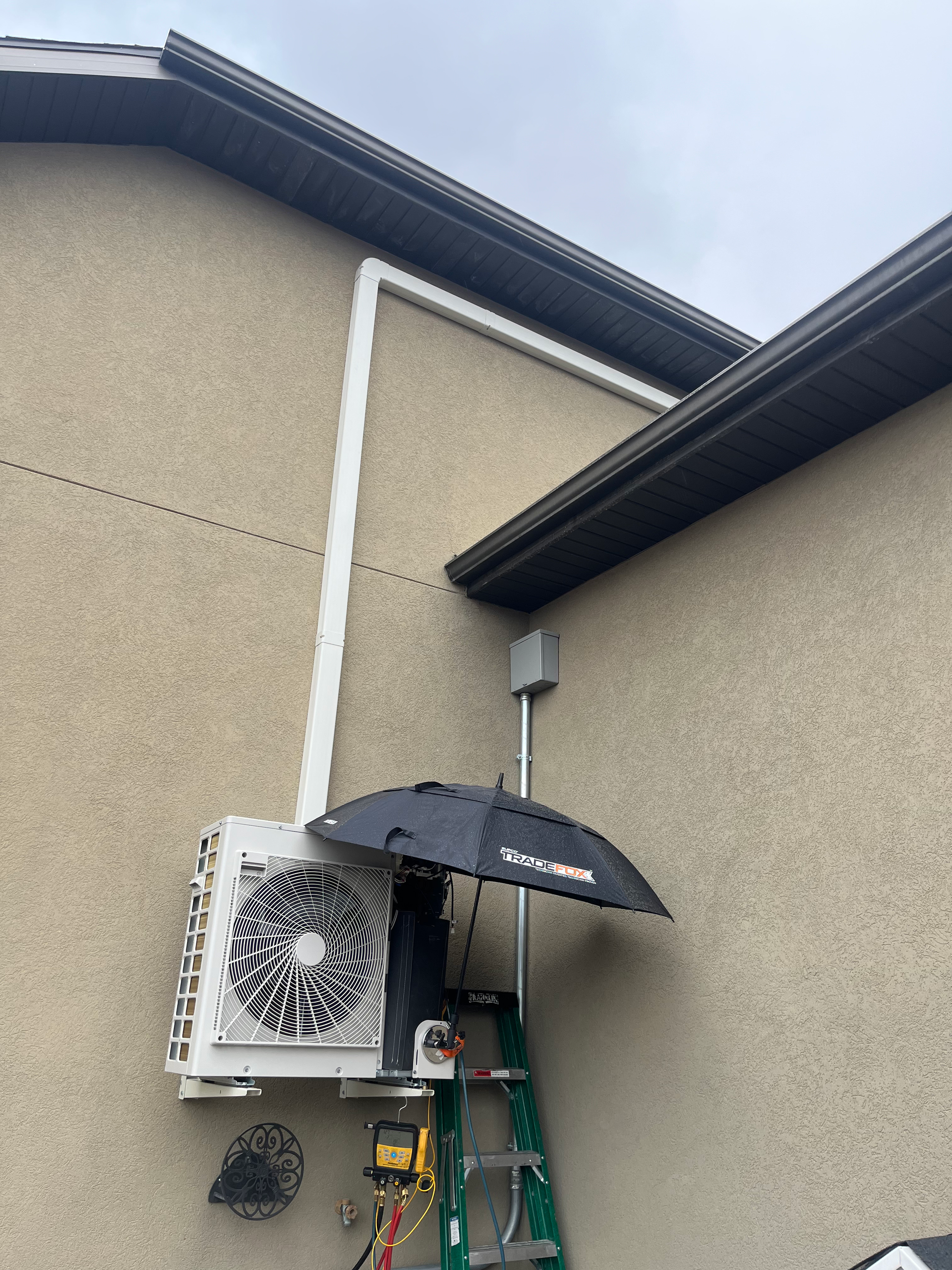 Person under umbrella on a ladder working on an HVAC unit, mounted on the side of a building.