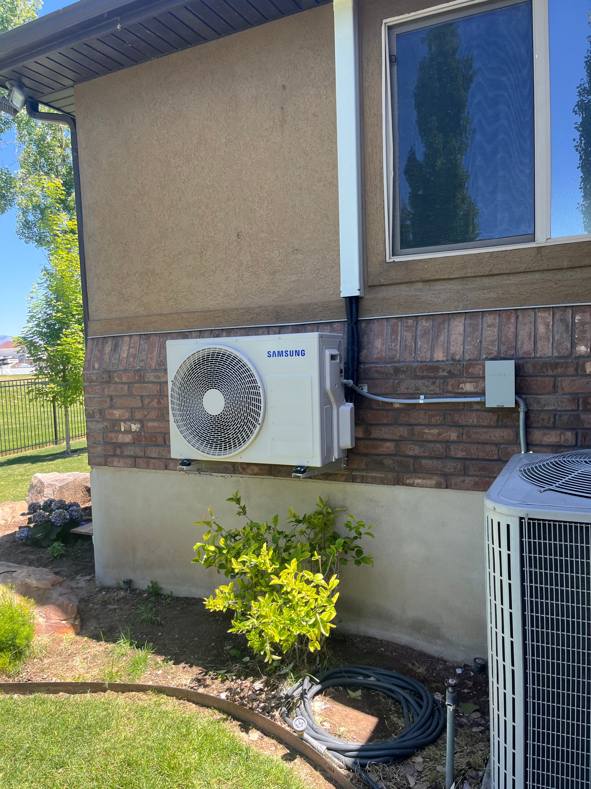 An air conditioning unit mounted on a brick wall next to a window and another unit on the ground.