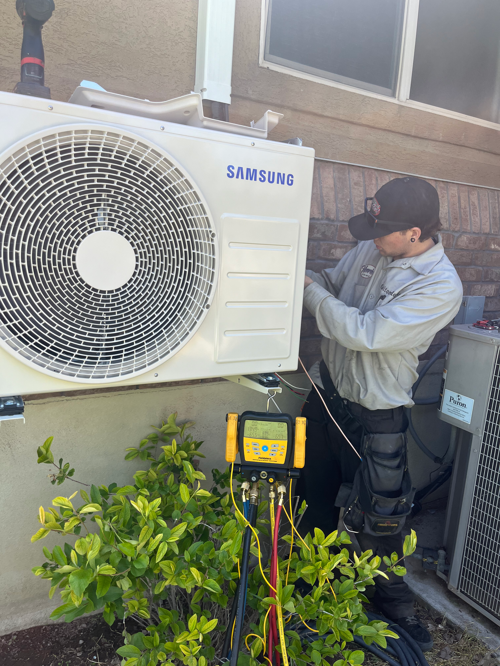 HVAC technician working on a Samsung air conditioning unit outside a building. He is wearing a cap.