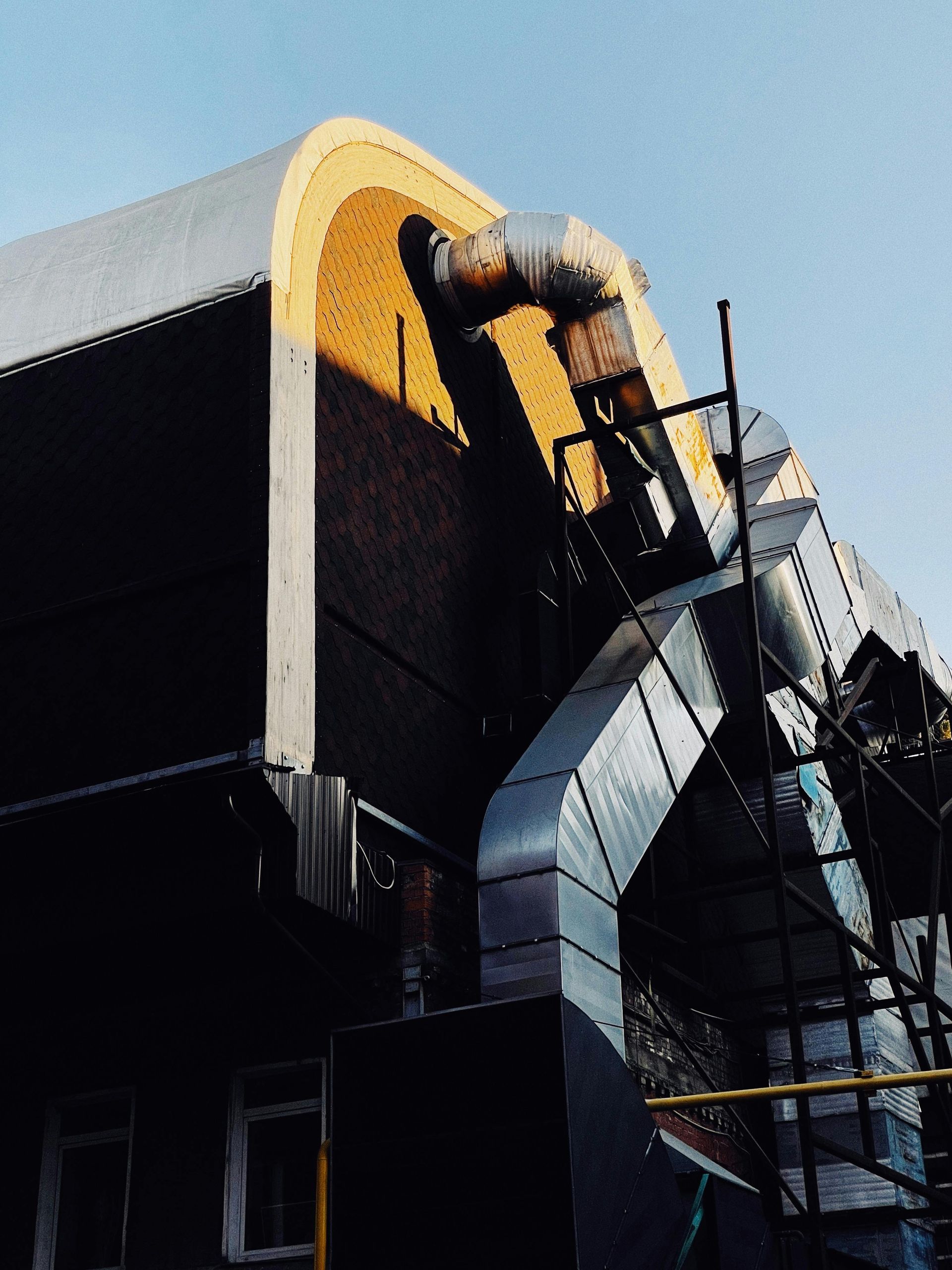 Building exterior with curved roof and metal ductwork, set against a blue sky.