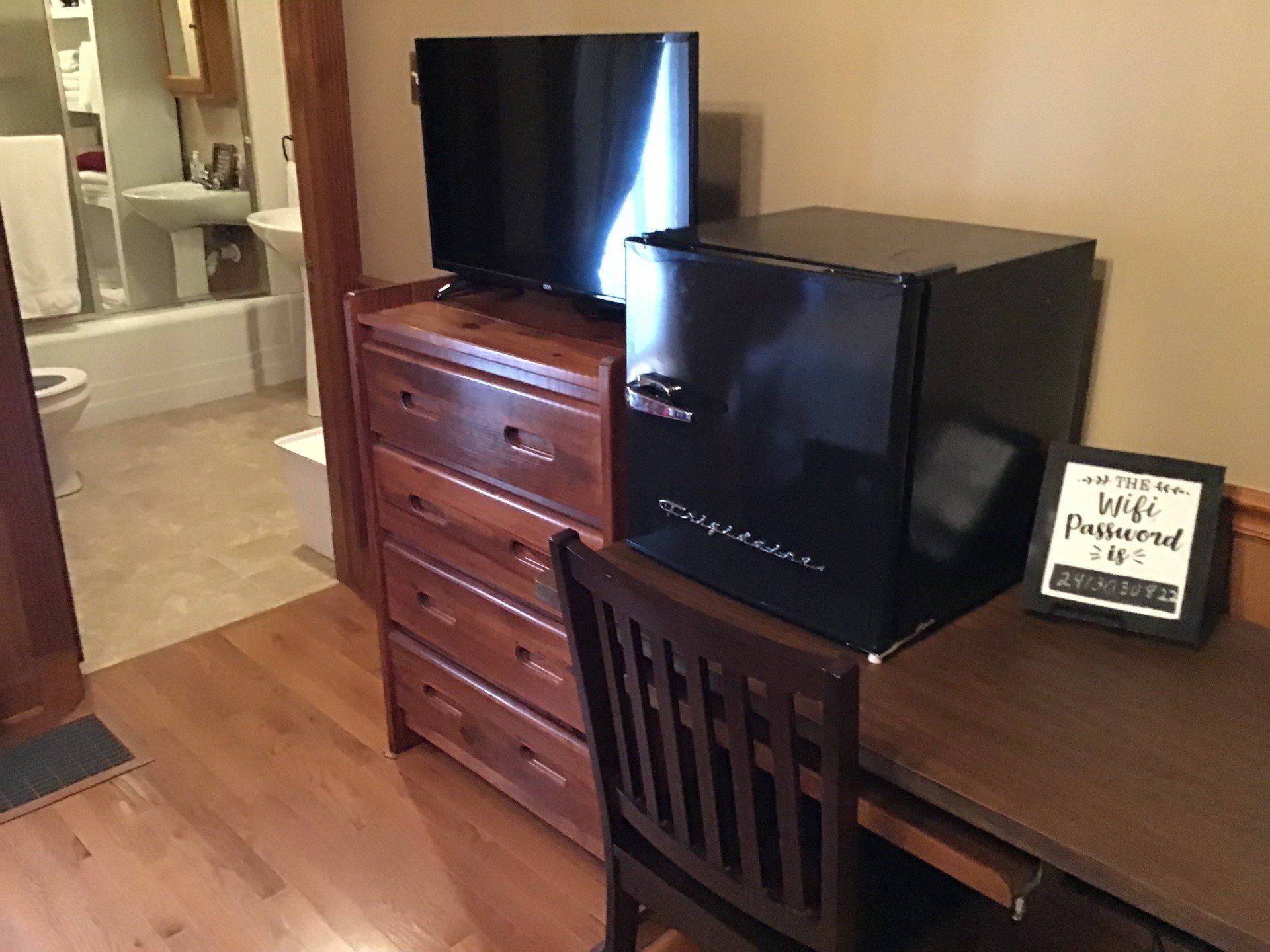 A room with a desk , chair , dresser , refrigerator and television.