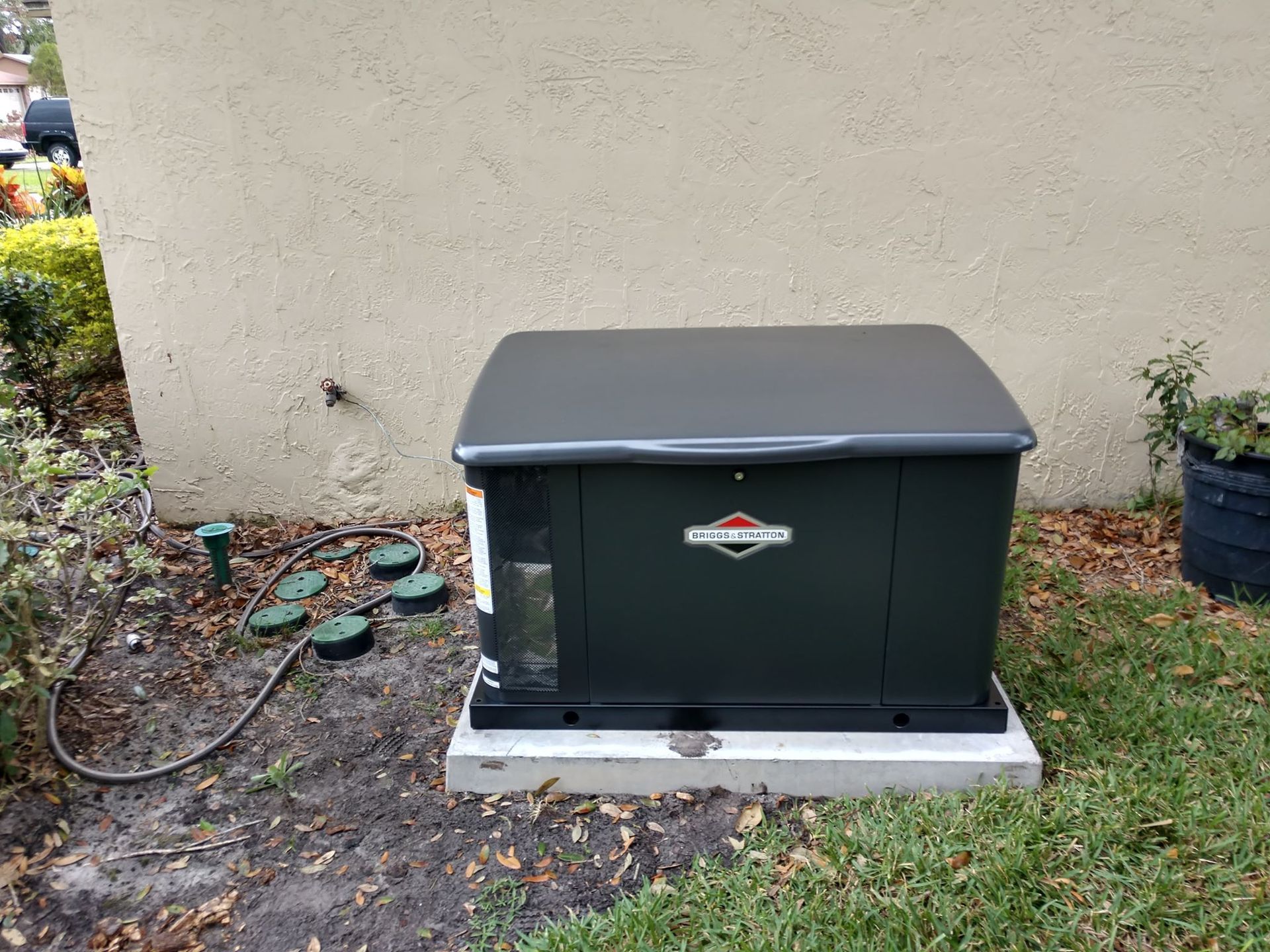Dark green Briggs & Stratton home generator on a concrete pad near a beige wall and grass.