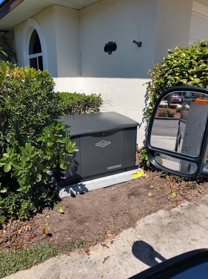 A dark generator box installed on a concrete base next to a house with bushes and a side mirror refl