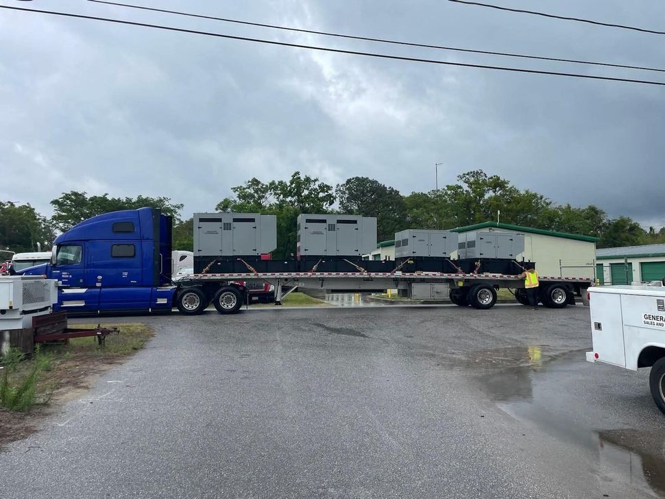 Blue semi-truck with flatbed trailer carrying four large generators under overcast sky.