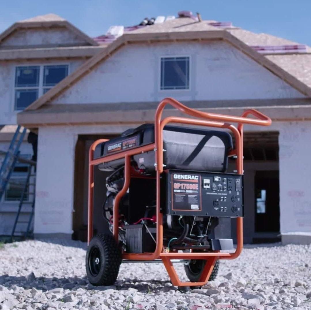A black generator on a brick base next to a white house with a meter and hose in the yard.