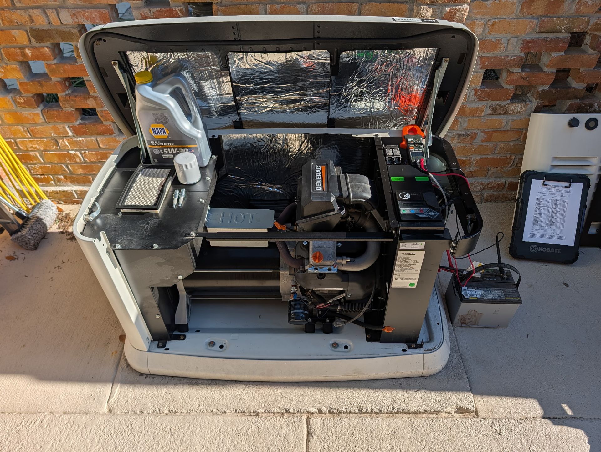A dark generator box sits on a concrete base next to a house with bushes, viewed from a vehicle mirror.