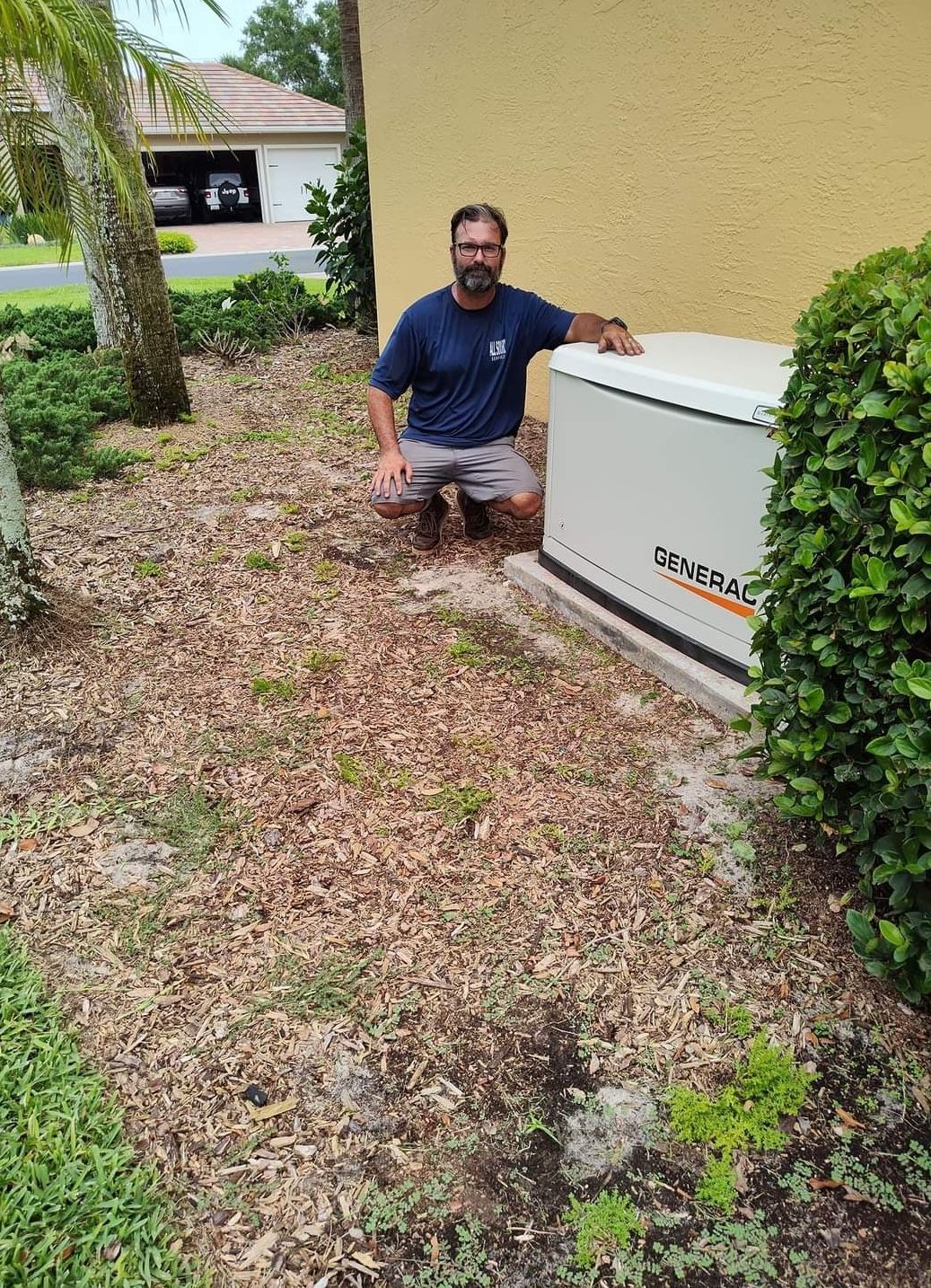 Man crouching next to a Generac generator against a yellow building with landscaping.