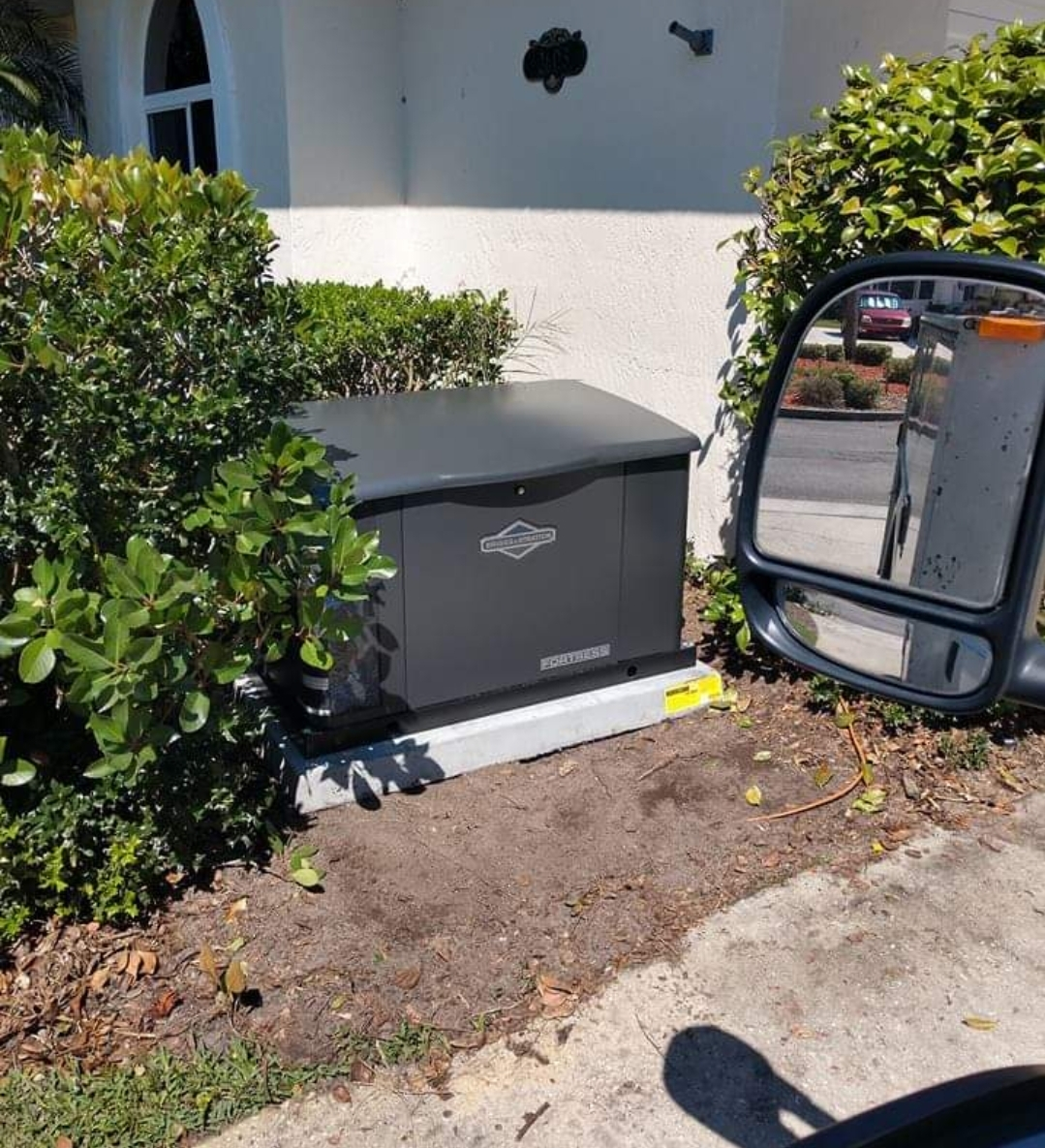 A dark generator box sits on a concrete base next to a house with bushes, viewed from a vehicle mirror.