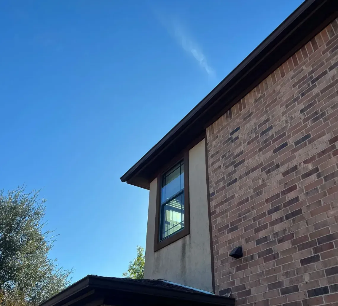 Brick house exterior with tan siding, dark roof trim, and a window against a blue sky