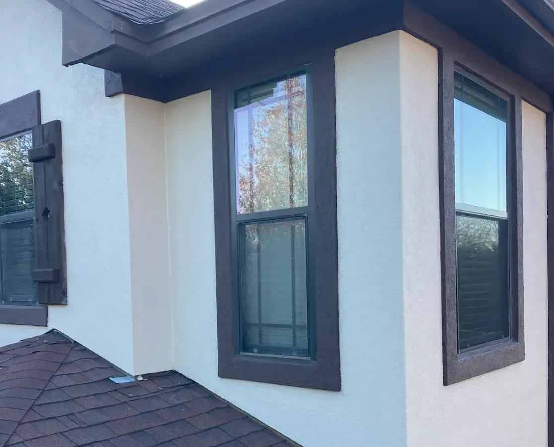 Exterior corner of a house with white stucco walls, dark trim, and tall windows above a shingled roof