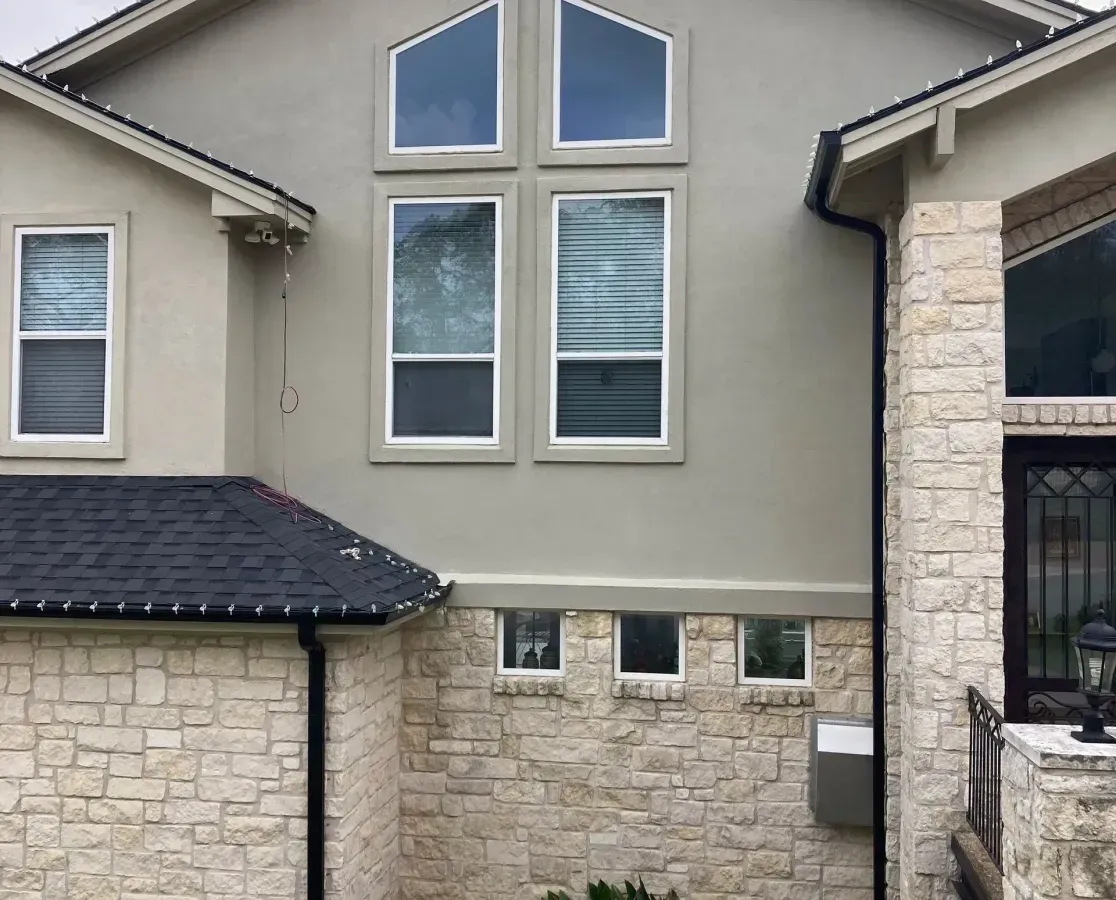 Modern beige stone-and-stucco house exterior with large windows and a small grassy front yard.