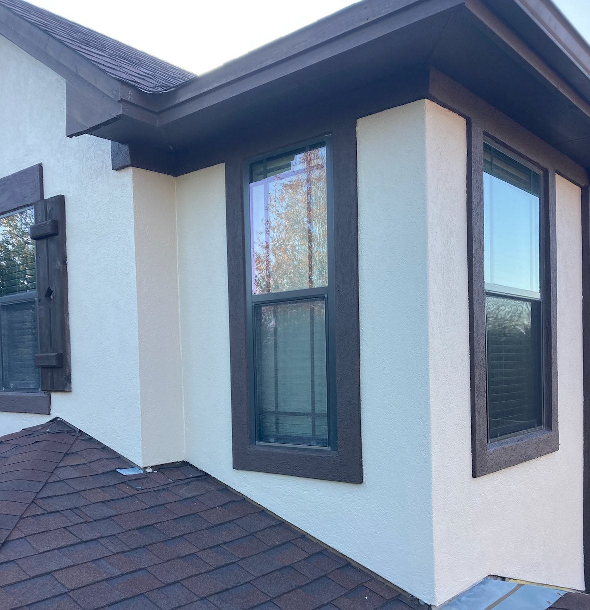 Exterior corner of a house with white stucco walls, dark trim, and tall windows above a shingled roof