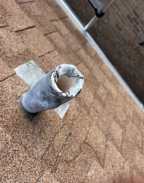 Damaged metal vent pipe protruding from a brown shingled roof under a brick wall.