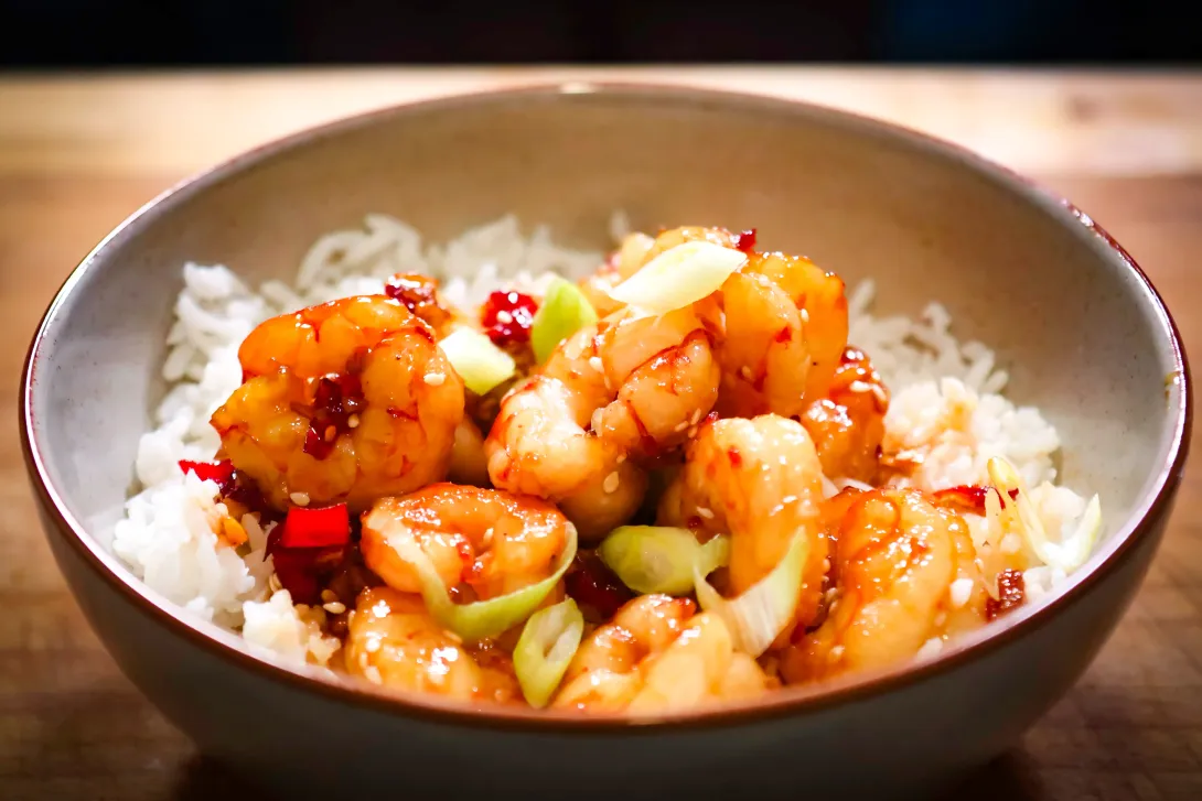 Cooked shrimp in orange sauce, held with chopsticks, in a black bowl.