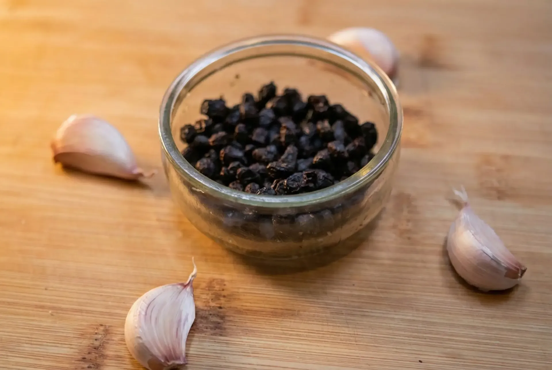 Small glass jar filled with black peppercorns surrounded by four garlic cloves on a wooden surface.