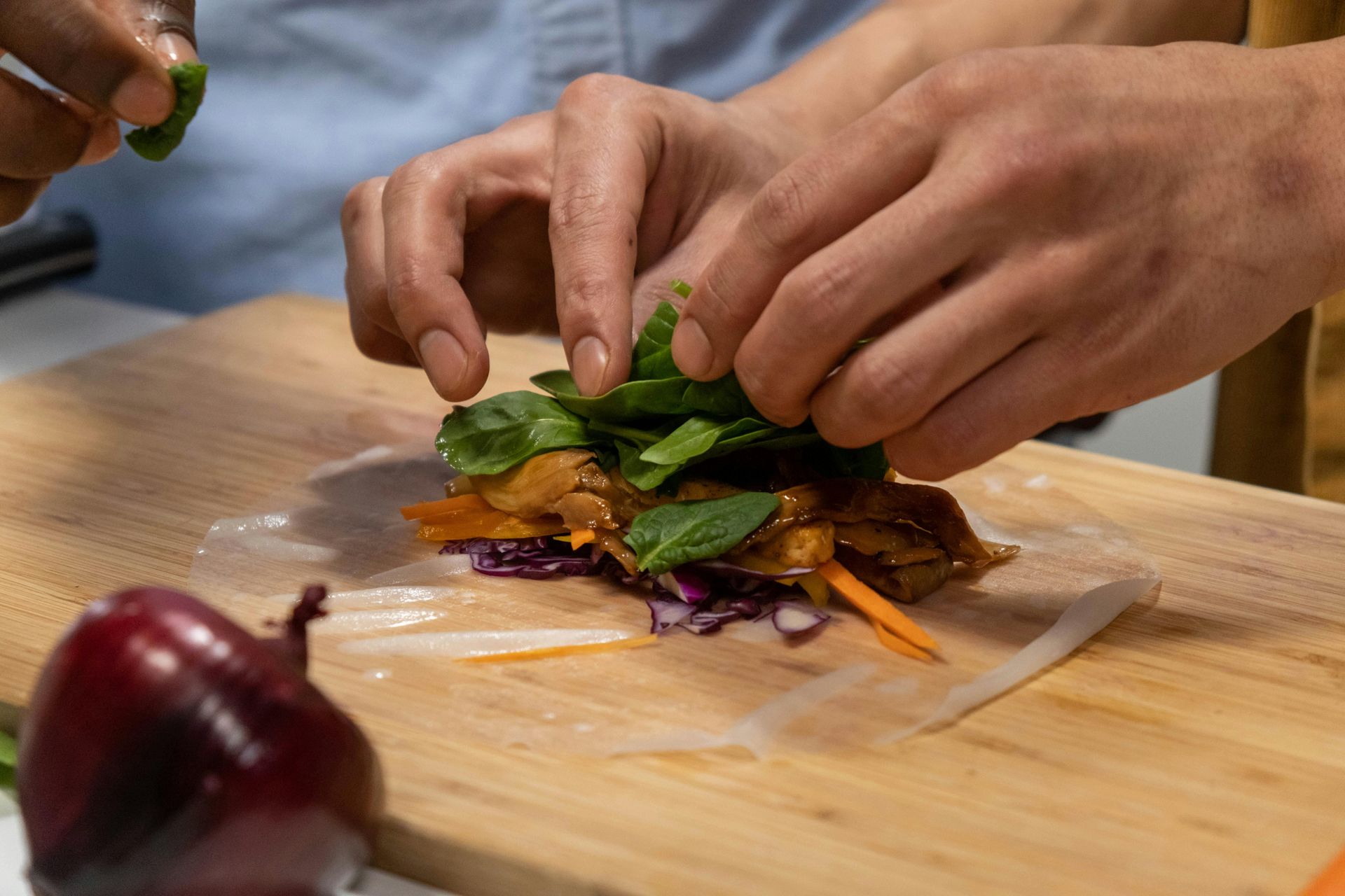Hands arranging spinach on a spring roll with vegetables on a wooden cutting board.